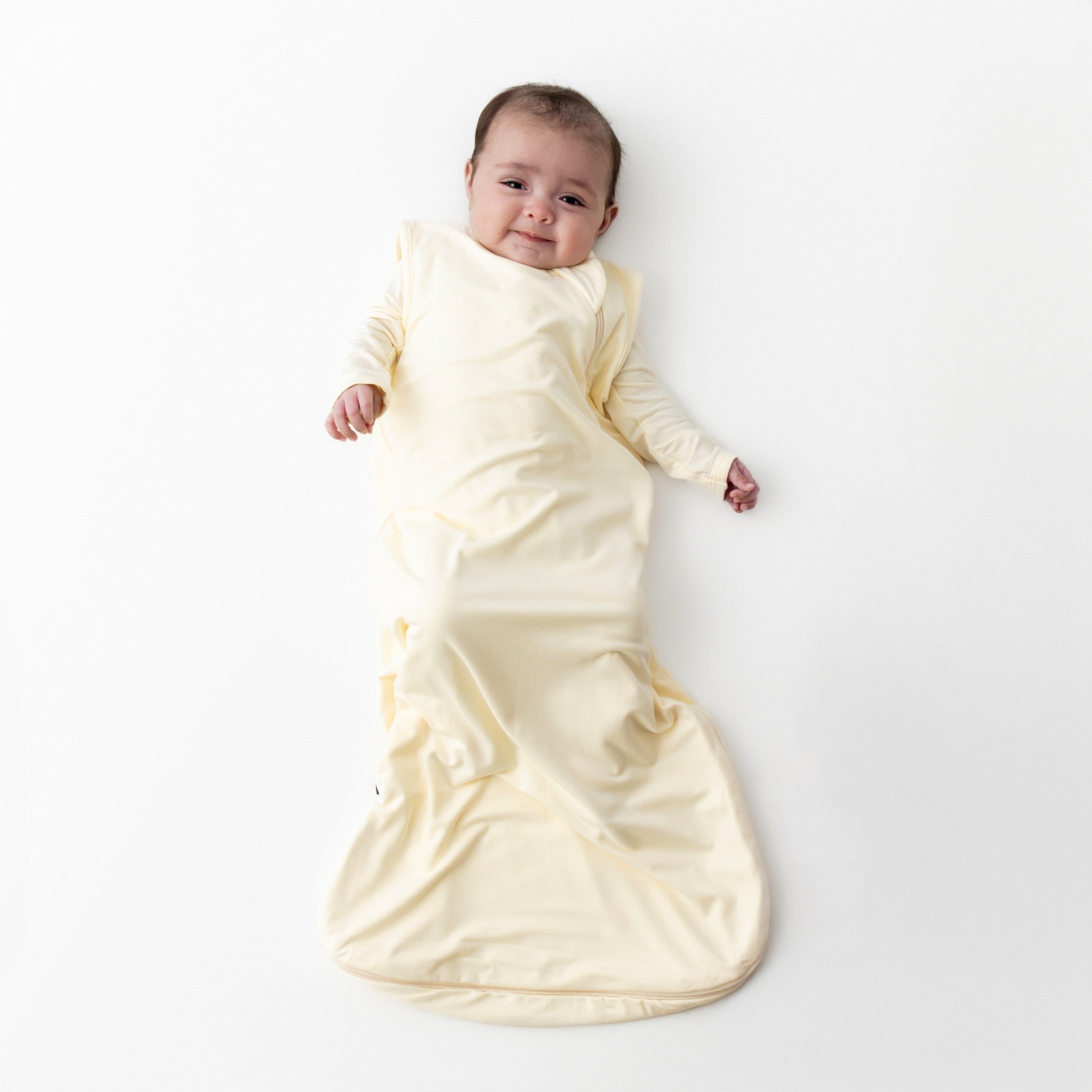 Smiling infant laying on a white surface wearing the soft and breathable Sleep Bag in Chamomile 0.5 with matching romper on underneath