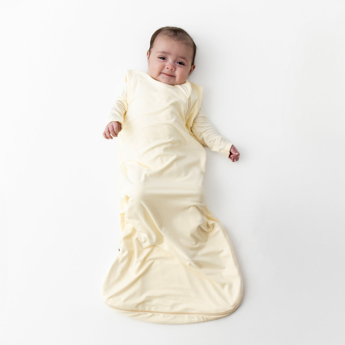 Smiling infant laying on a white surface wearing the soft and breathable Sleep Bag in Chamomile 0.5 with matching romper on underneath