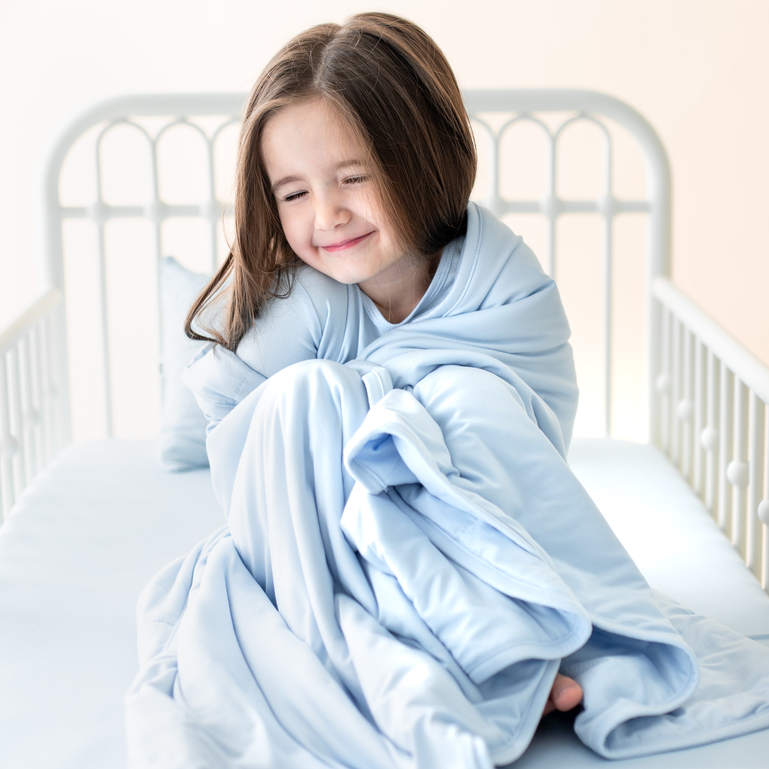 Young girl sitting in bed cuddling the Toddler Blanket in Breeze 1.0 around her shoulders