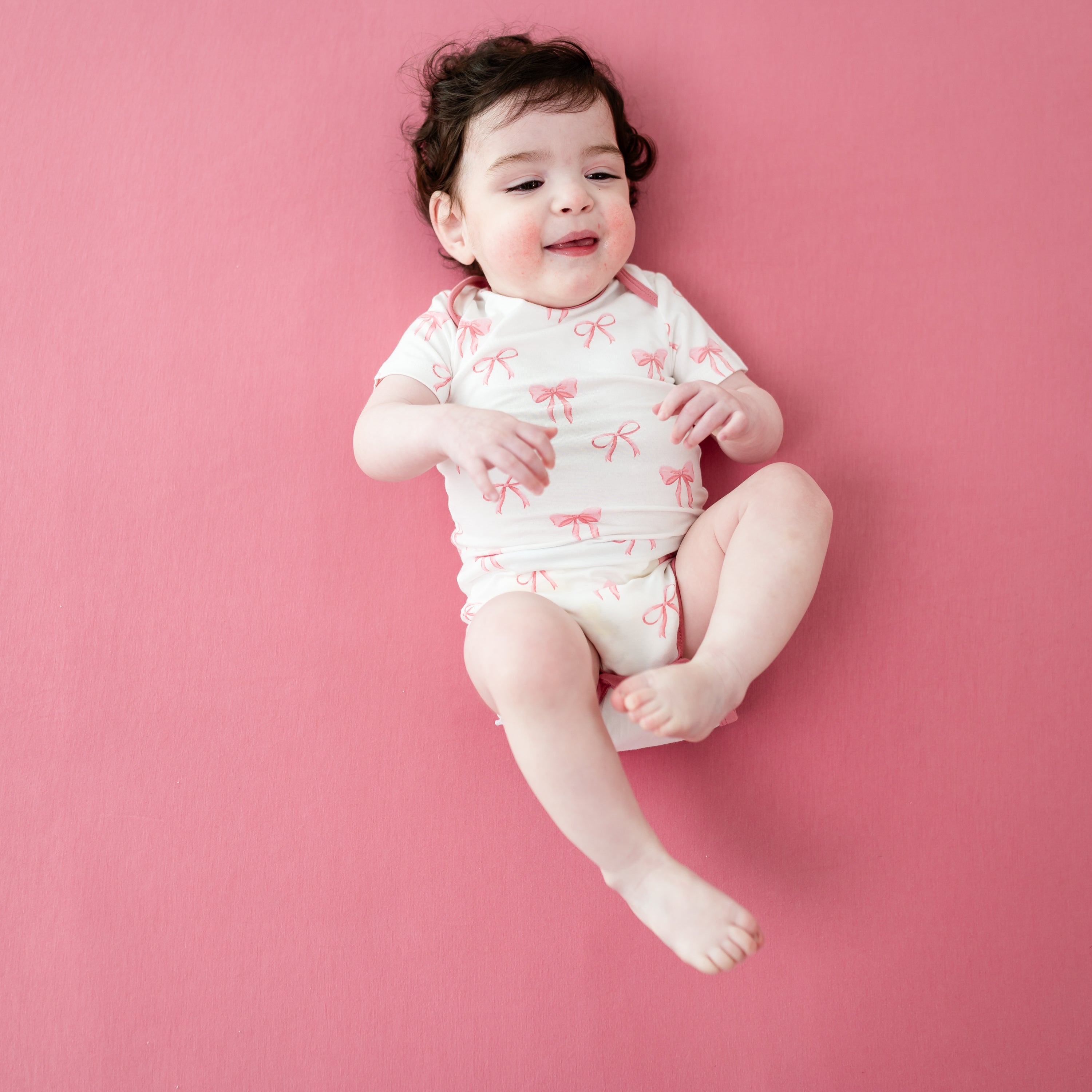 Young toddler laying down on an apple blossom crib sheet wearing Bodysuit in Bow