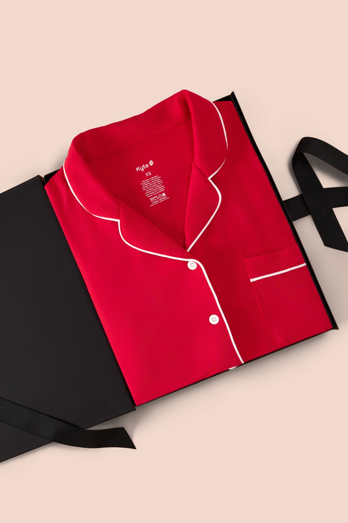 A folded red pajama shirt with white piping is presented in a black gift box on a light beige background. The pajama top has a classic collar, button front, and a chest pocket with matching white piping.