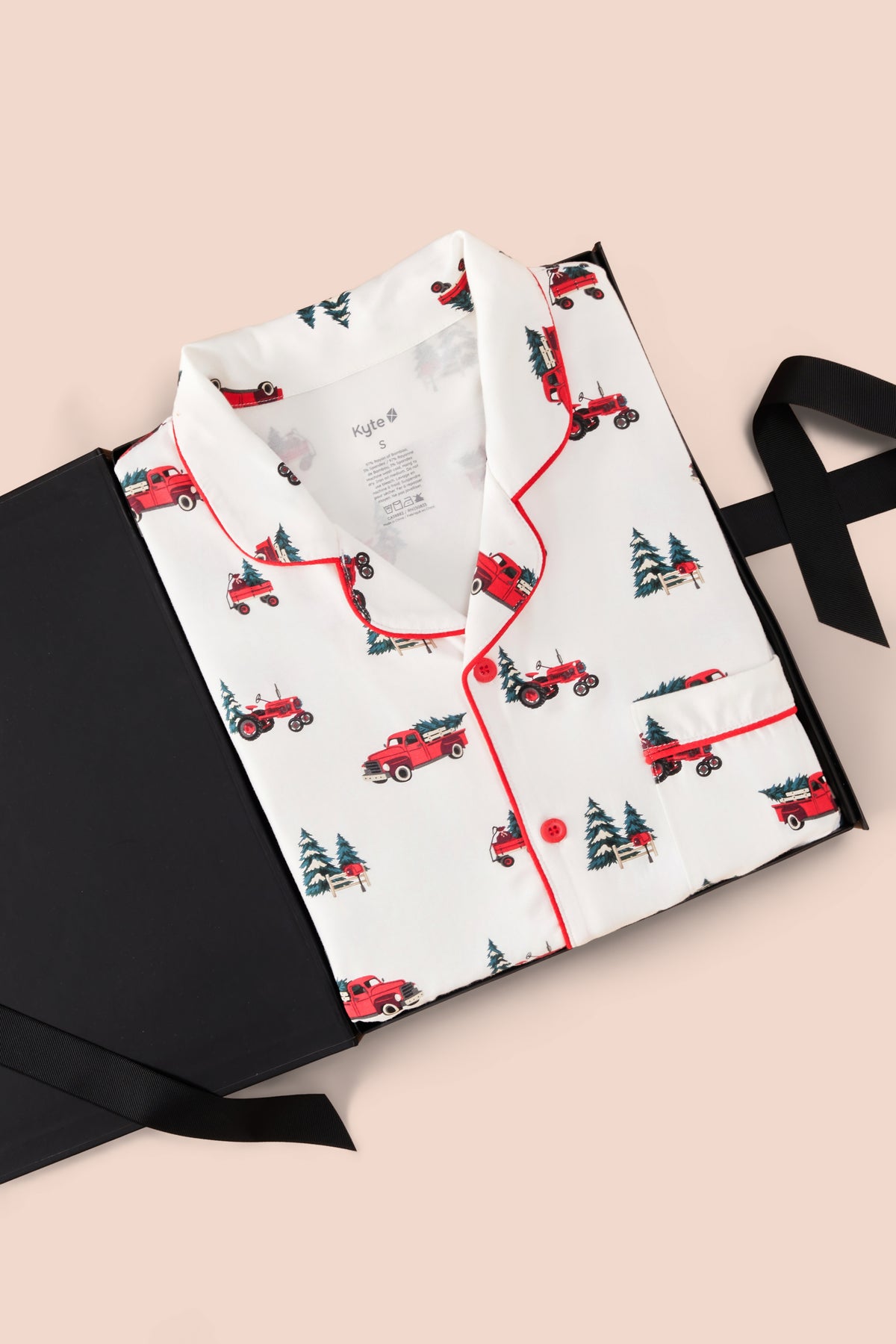 A folded white pajama shirt with red piping and a red Christmas tree tractor print, presented on a black gift box with a black ribbon against a light pink background.
