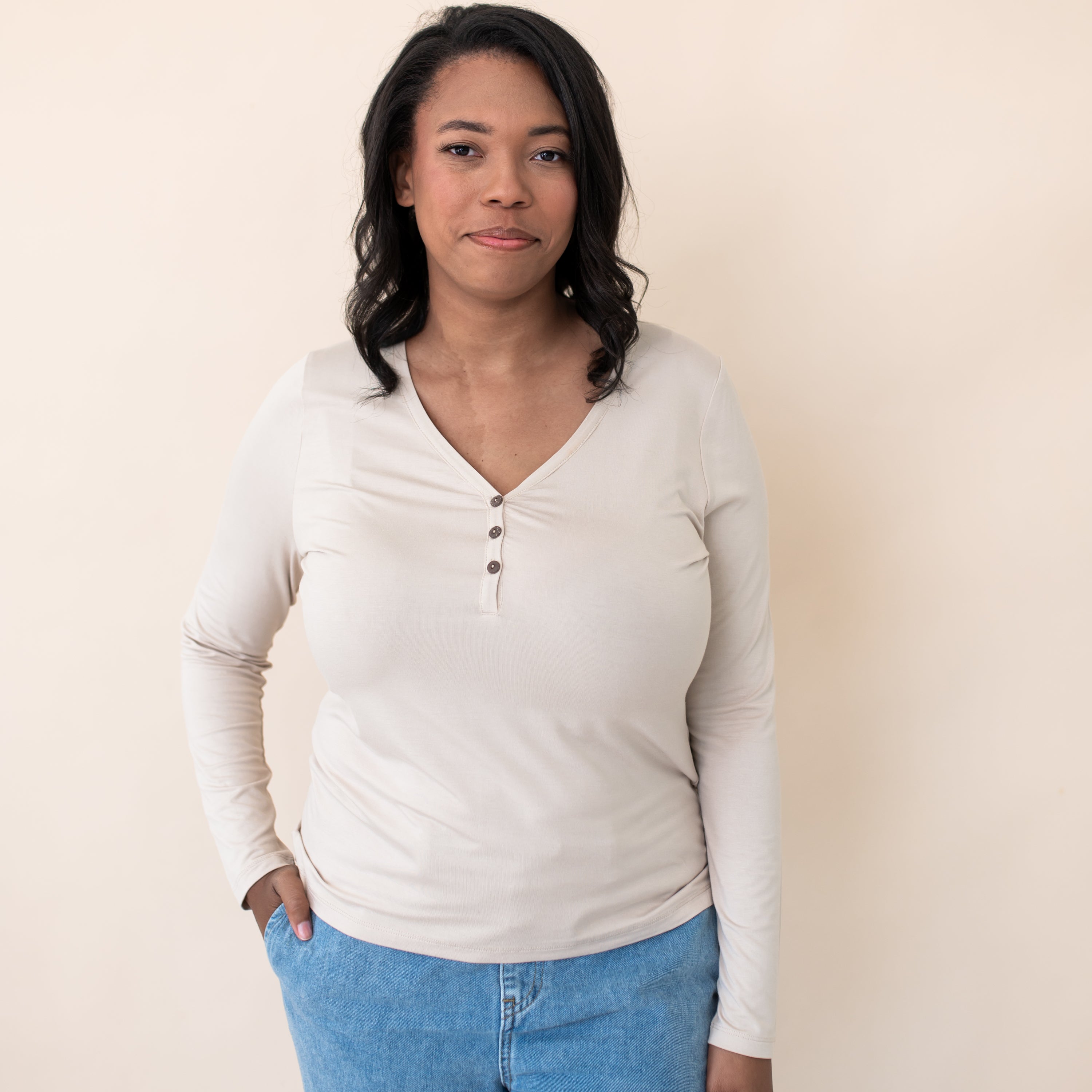 Smiling female model wearing the Women's Long Sleeve Henley Tee in Bisque with jeans standing in front of a beige backdrop