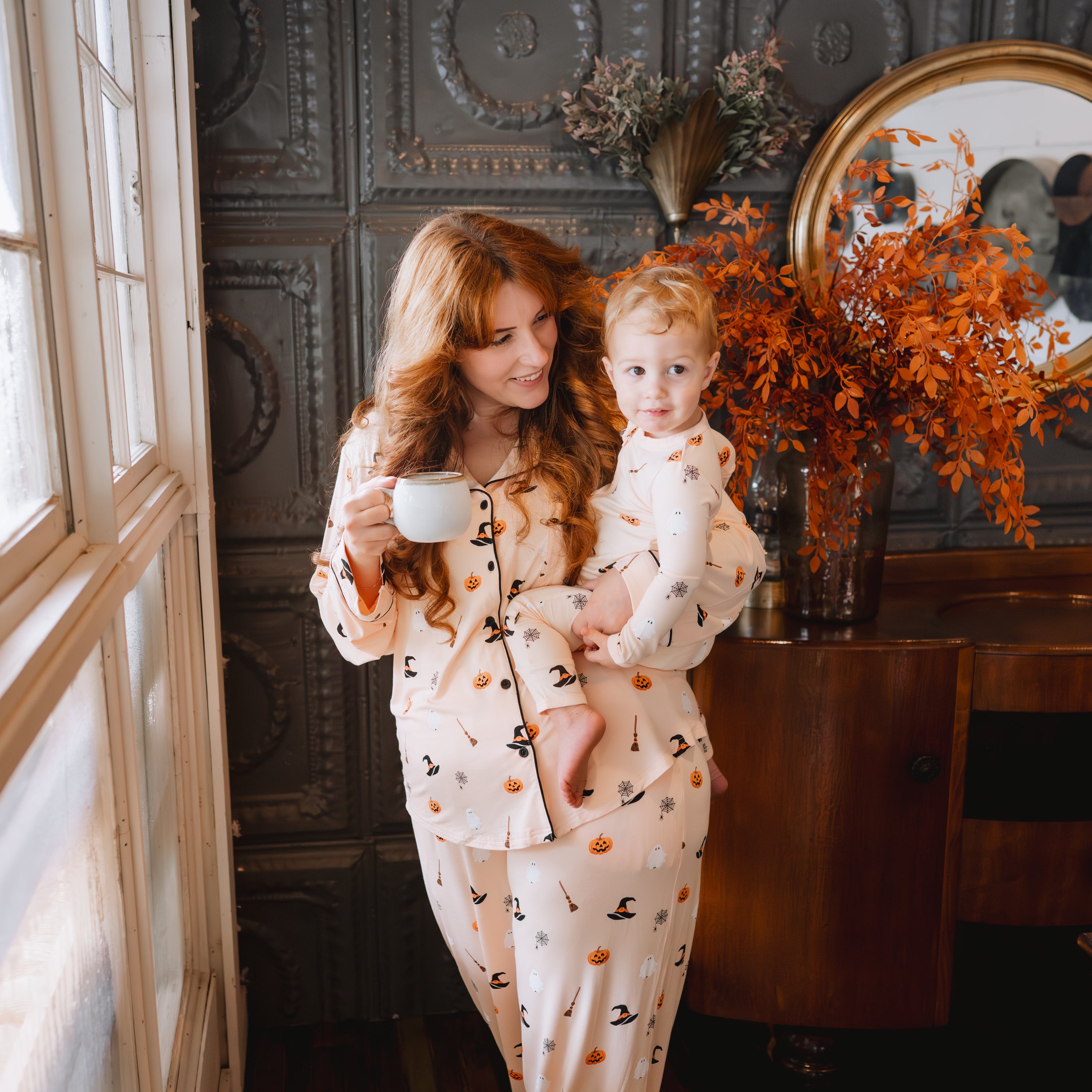 Mother wearing the Long-Sleeved Women's Pajama Set in Bewitched holding her son who is in a matching zippered romper and holding a cup of coffee in the other hand