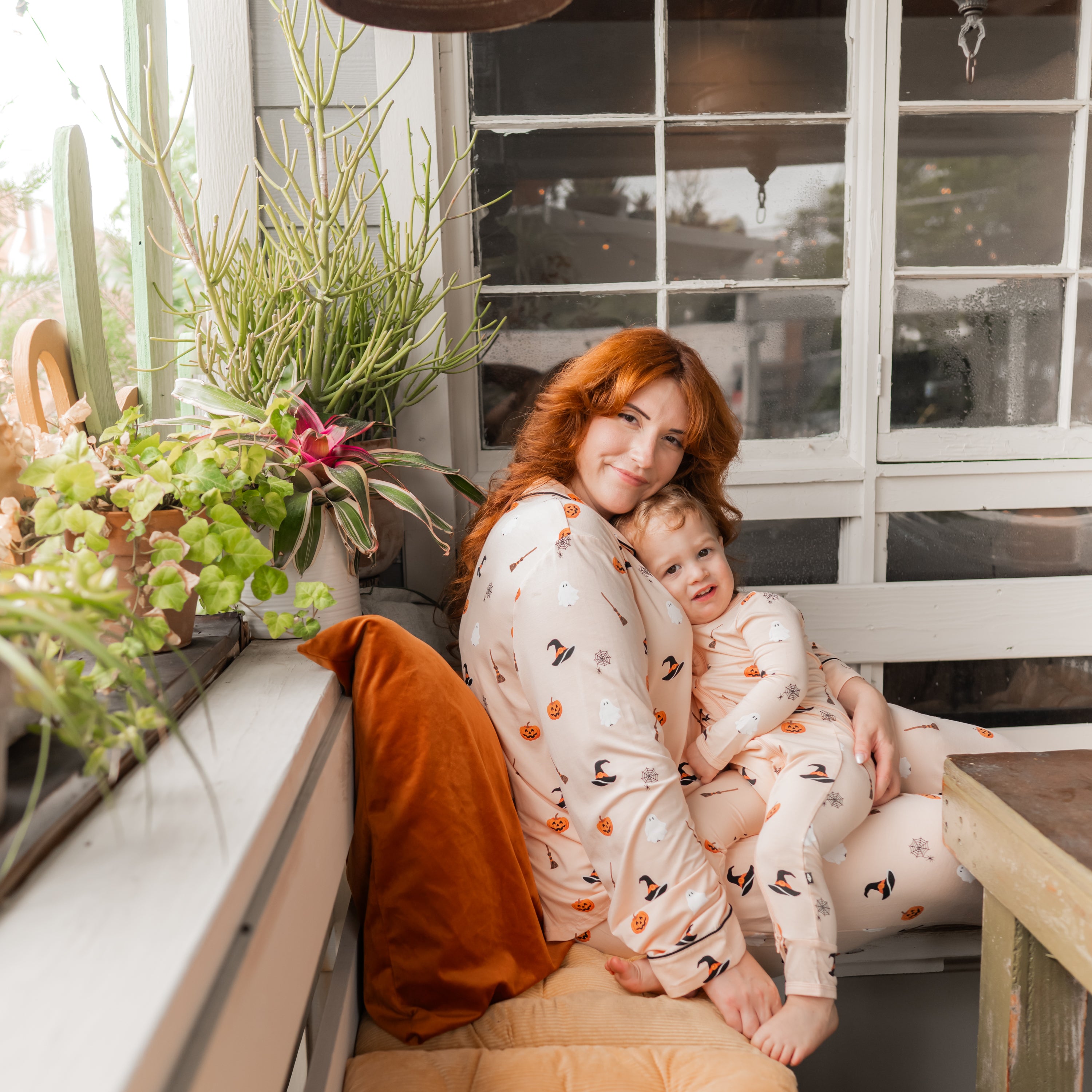 Mother wearing the Long-Sleeved Women's Pajama Set in Bewitched sitting on a bench with her son on her lap who is wearing a matching zippered romper