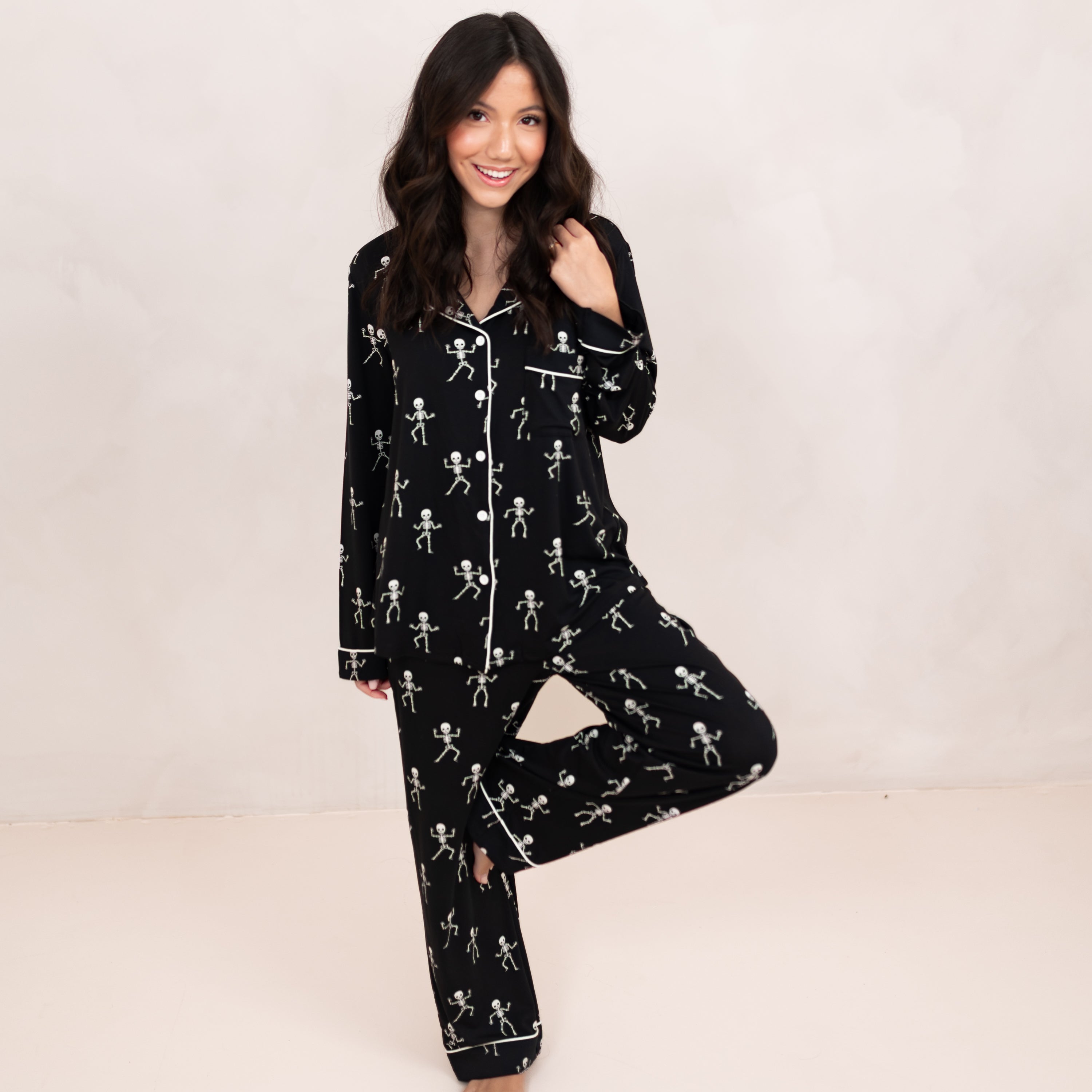 Smiling model standing on one leg with the other leg bent and resting on her knee wearing the Long-Sleeved Women's Pajama Set in Skeleton in front of a light beige backdrop