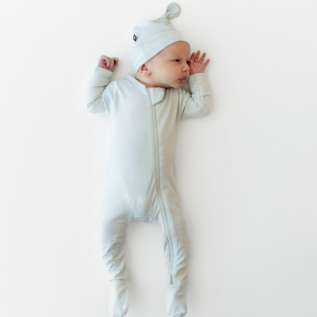 Newborn laying down with hands at their head wearing the Zippered Footie in Dew with matching knotted hat
