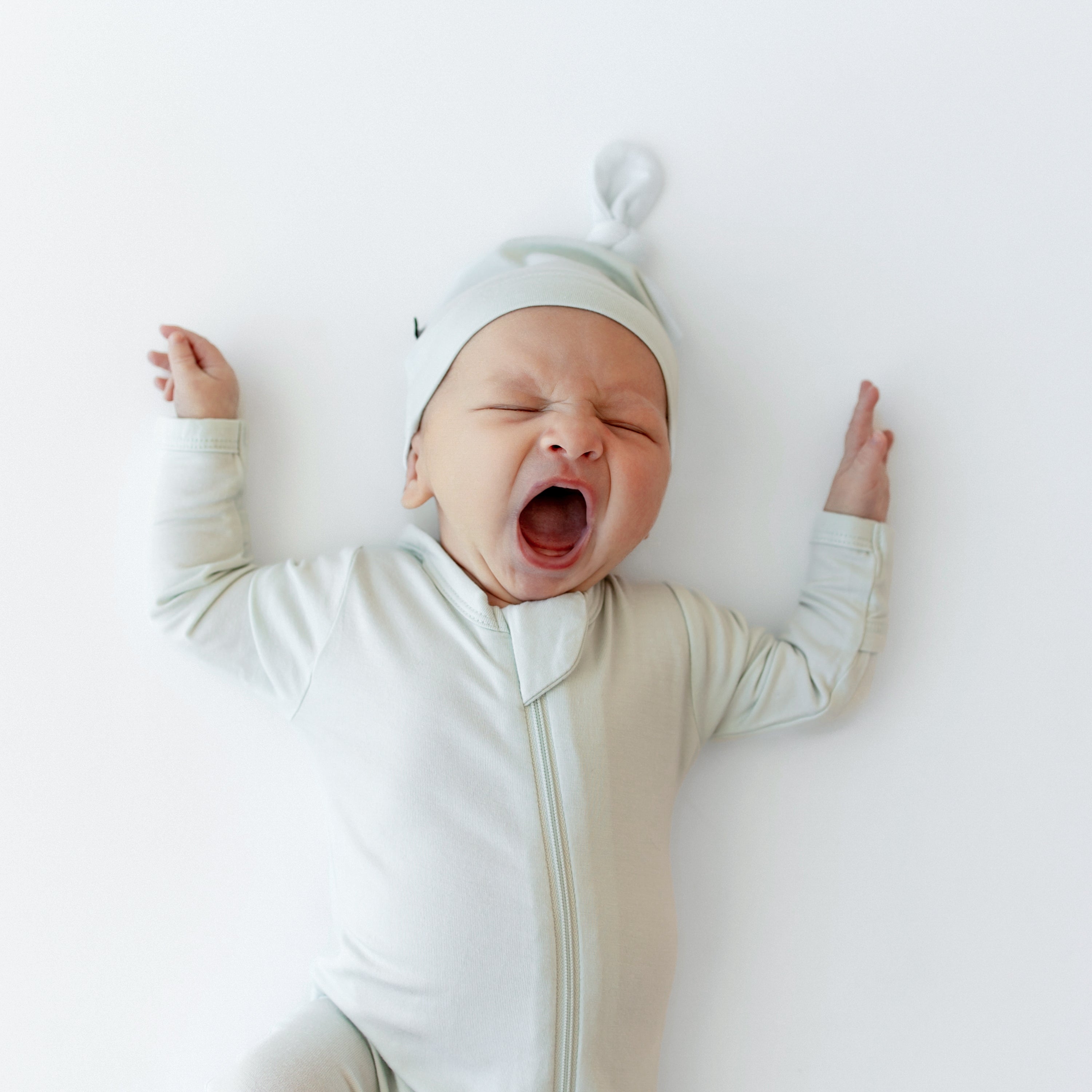 Close up of yawning newborn wearing the Zippered Footie in Dew with matching knotted hat