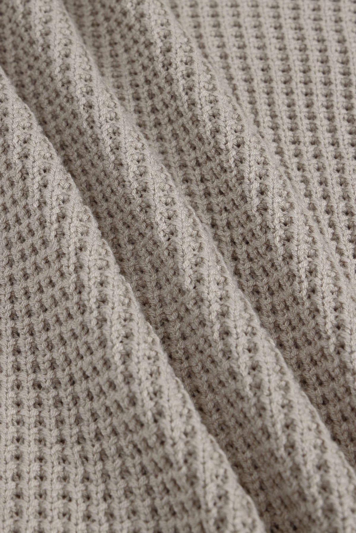 Close-up of a textured knit fabric with a waffle or basketweave pattern in a warm taupe-beige color, shown with soft folds.
