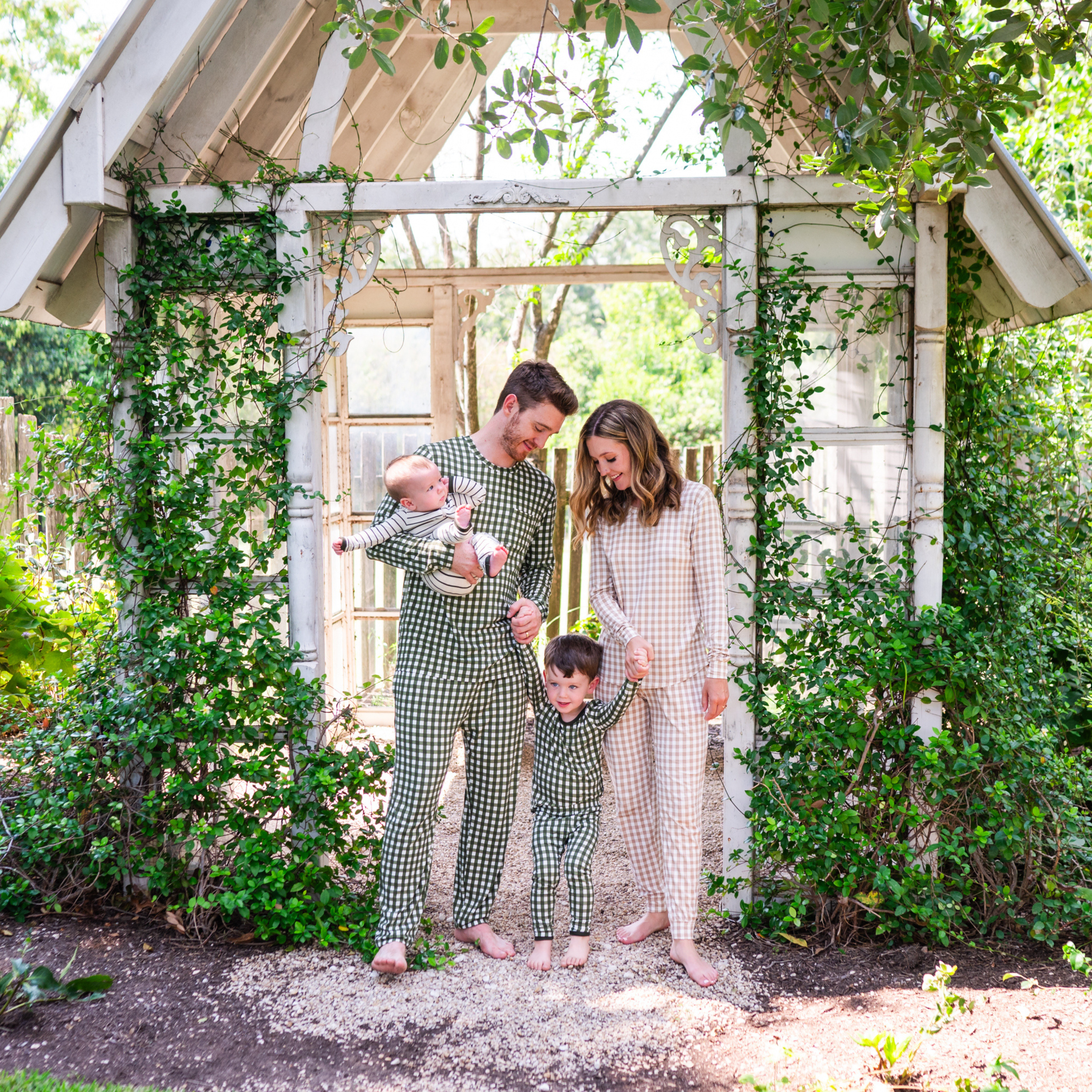Family of 4 standing in front of a white entry way with greenery around them. Mother wearing the Women's Jogger Pajama Set in Gingham Bisque, father and son matching in Gingham Fir pajamas with the father holding the youngest wearing a ribbed Fir Stripe Romper