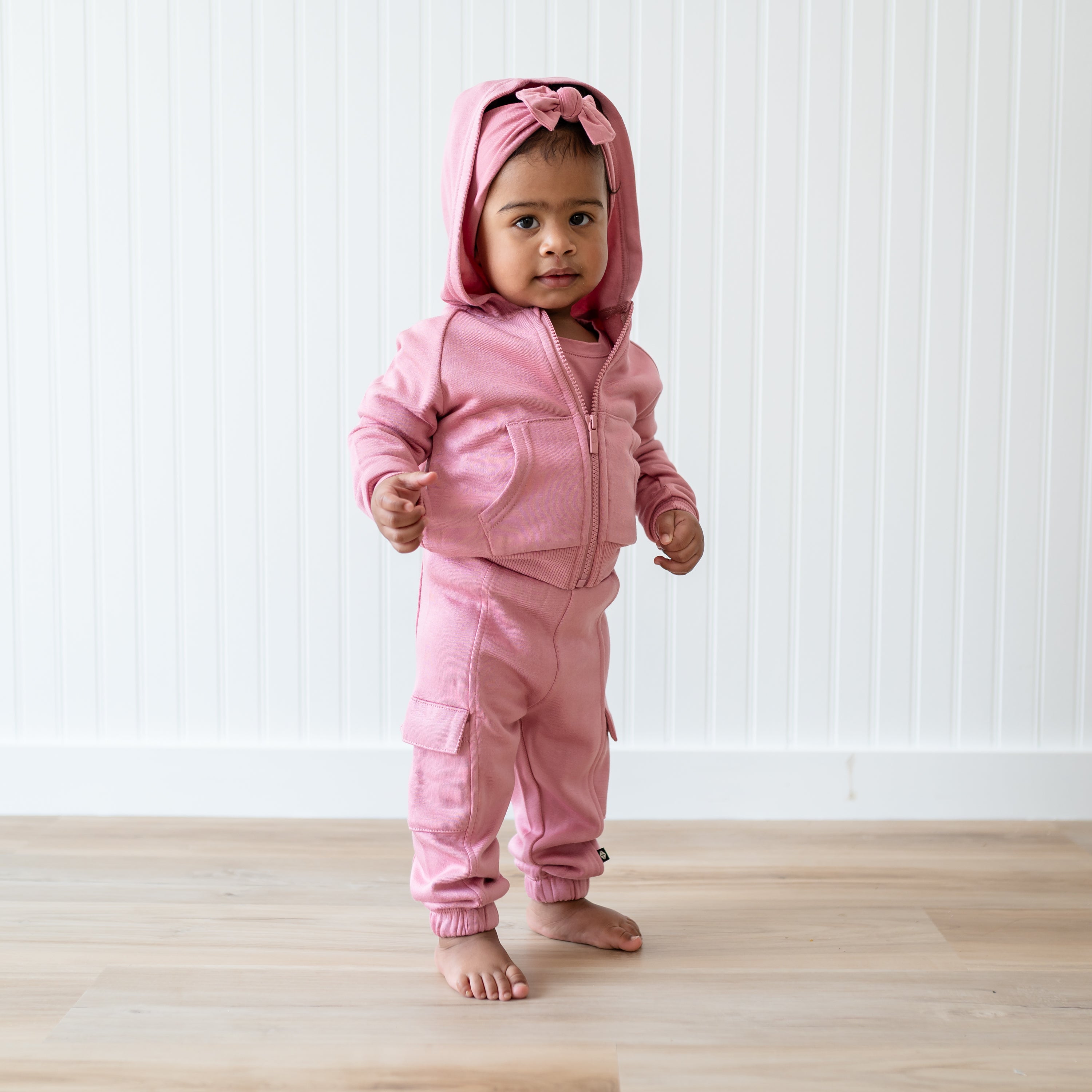 Young toddler standing wearing the Fleece Zip Up Hoodie in Apple Blossom with the hood up and matching Fleece Cargo pant standing in front of a white paneled wall