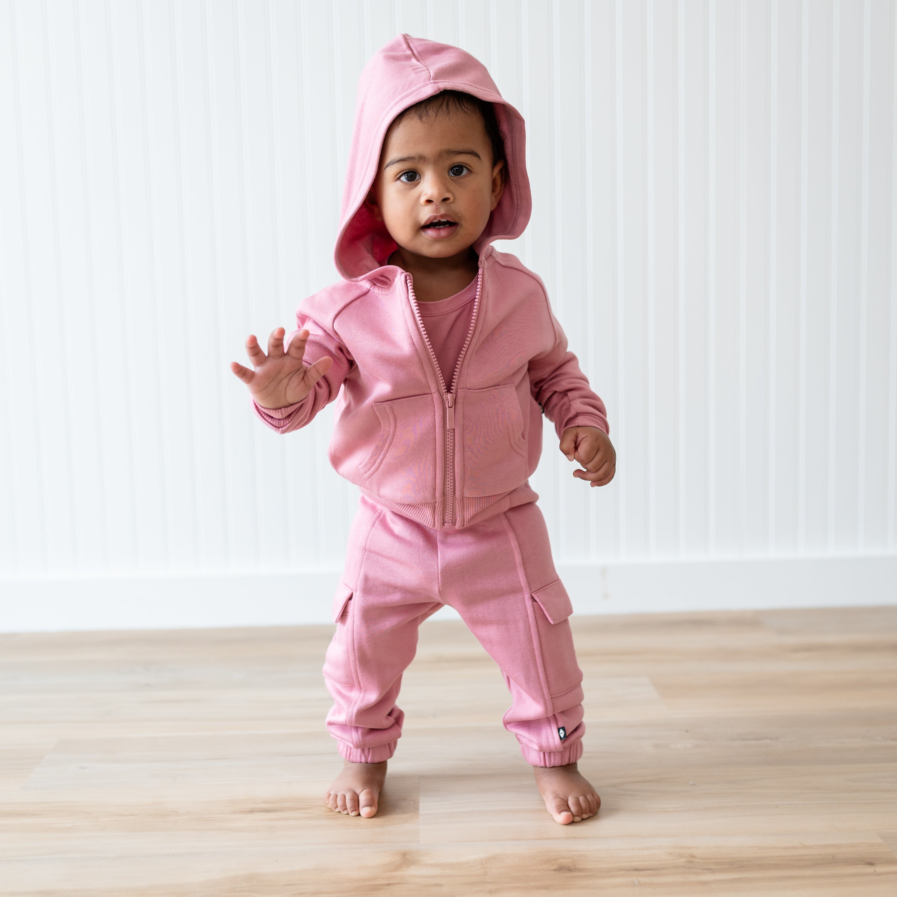 Young toddler walking wearing the Fleece Zip Up Hoodie in Apple Blossom with the hood up and the matching fleece cargo pants