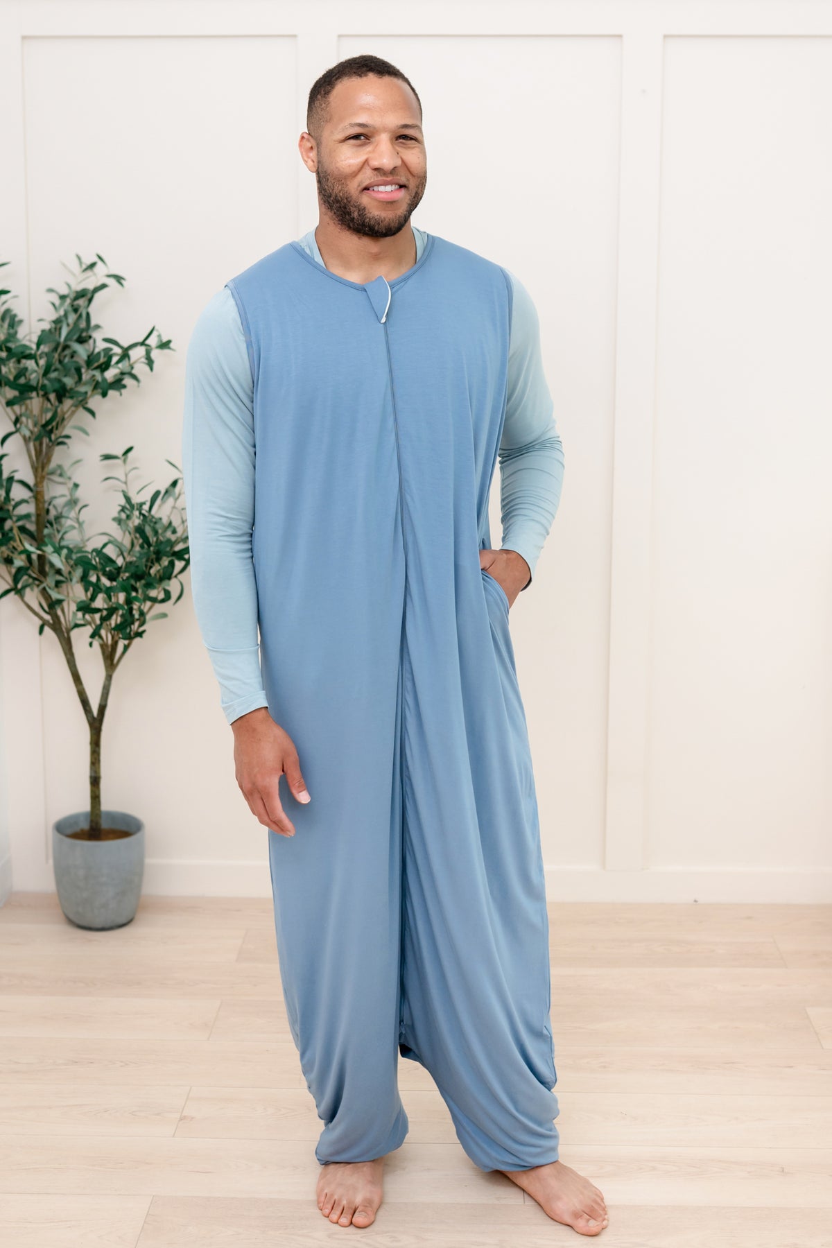 Man wearing a blue onesie standing in a room with a plant.