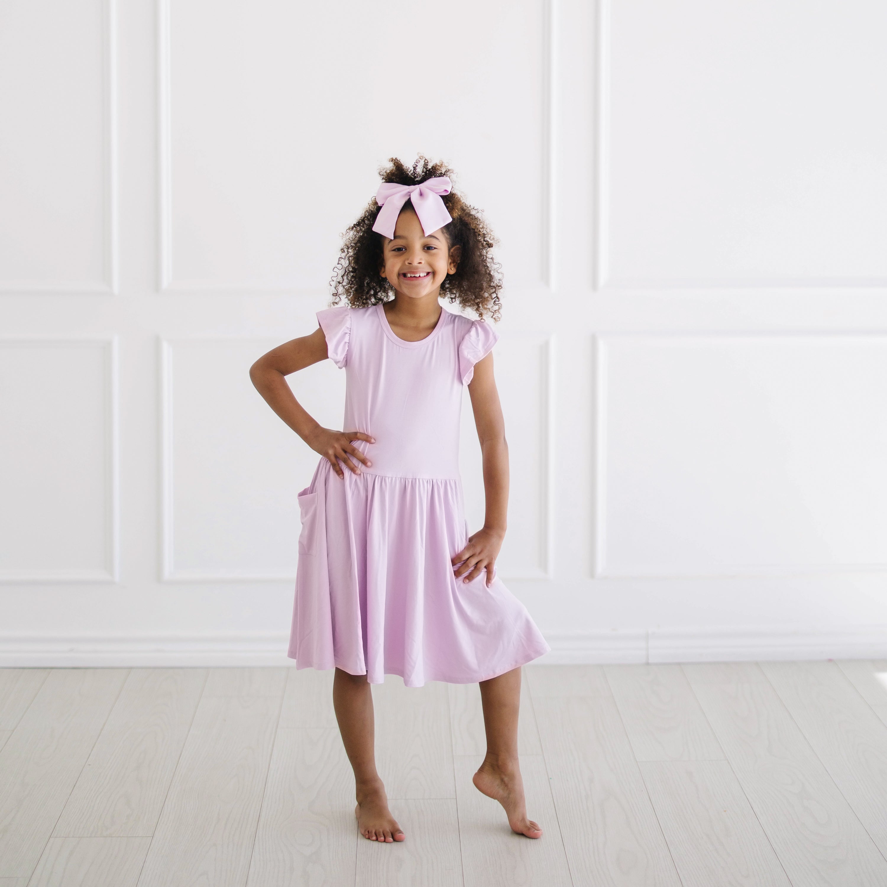 Young girl posing in front of a white wall wearing the Pocket Dress in Thistle with matching large alligator bow clip