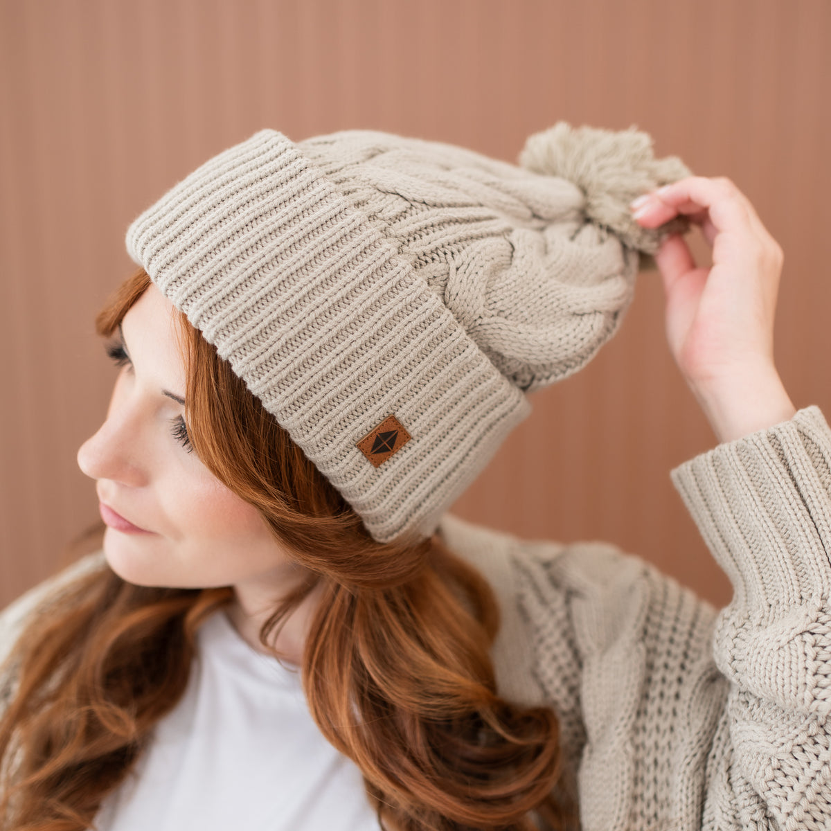 Cable Knit Adult Pom Beanie in Almond with the Kyte Baby vegan leather tag visible shown on a female model