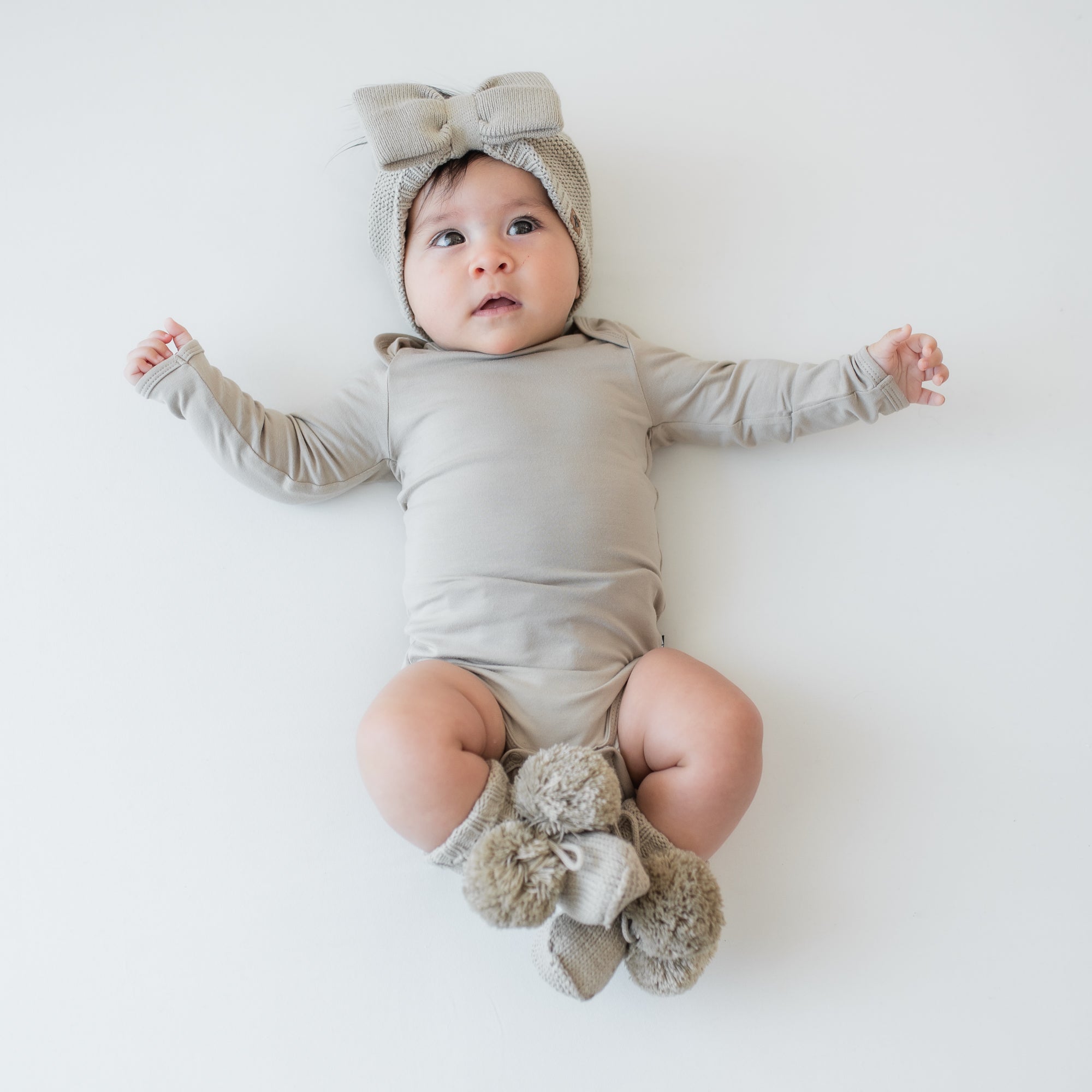 Infant laying down wearing the Chunky Knit Bow Headband in Almond paired with matching long sleeve bodysuit and knit booties