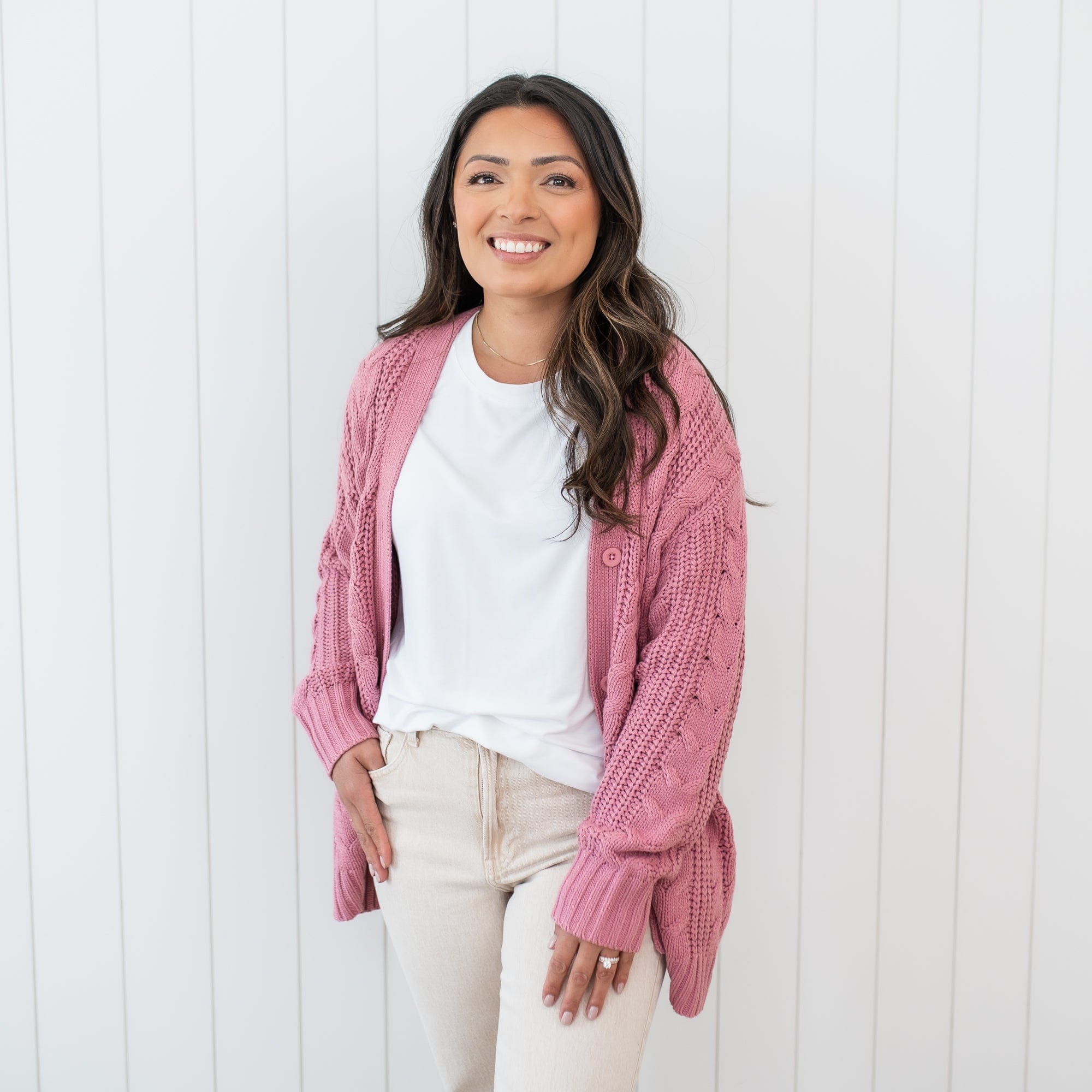 Female wearing the Cable Knit Adult Cardigan in Apple Blossom paired with a white shirt and beige colored pants