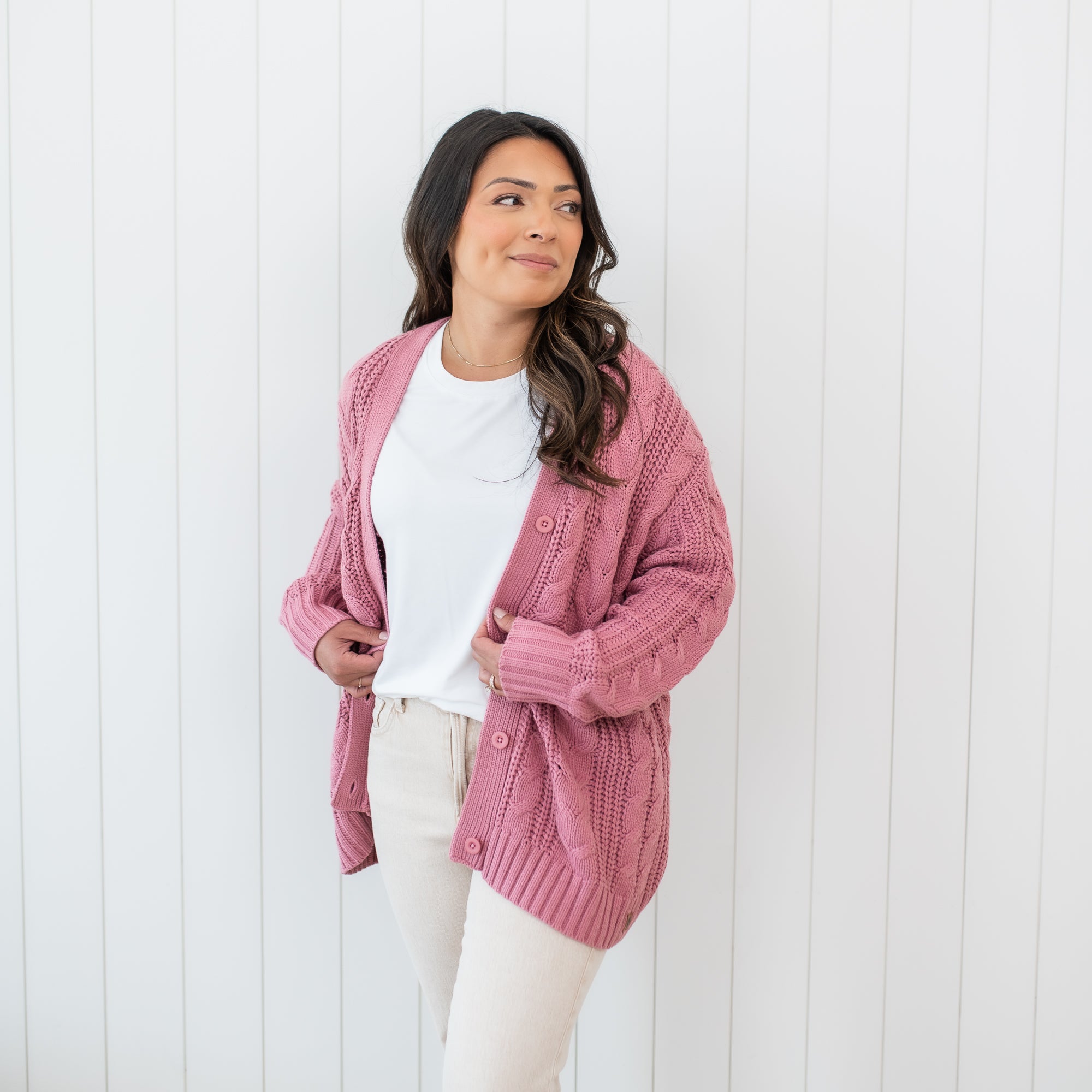 Smiling female model wearing the Cable Knit Adult Cardigan in Apple Blossom paired with cream pants and a white shirt standing in front of a white paneled wall