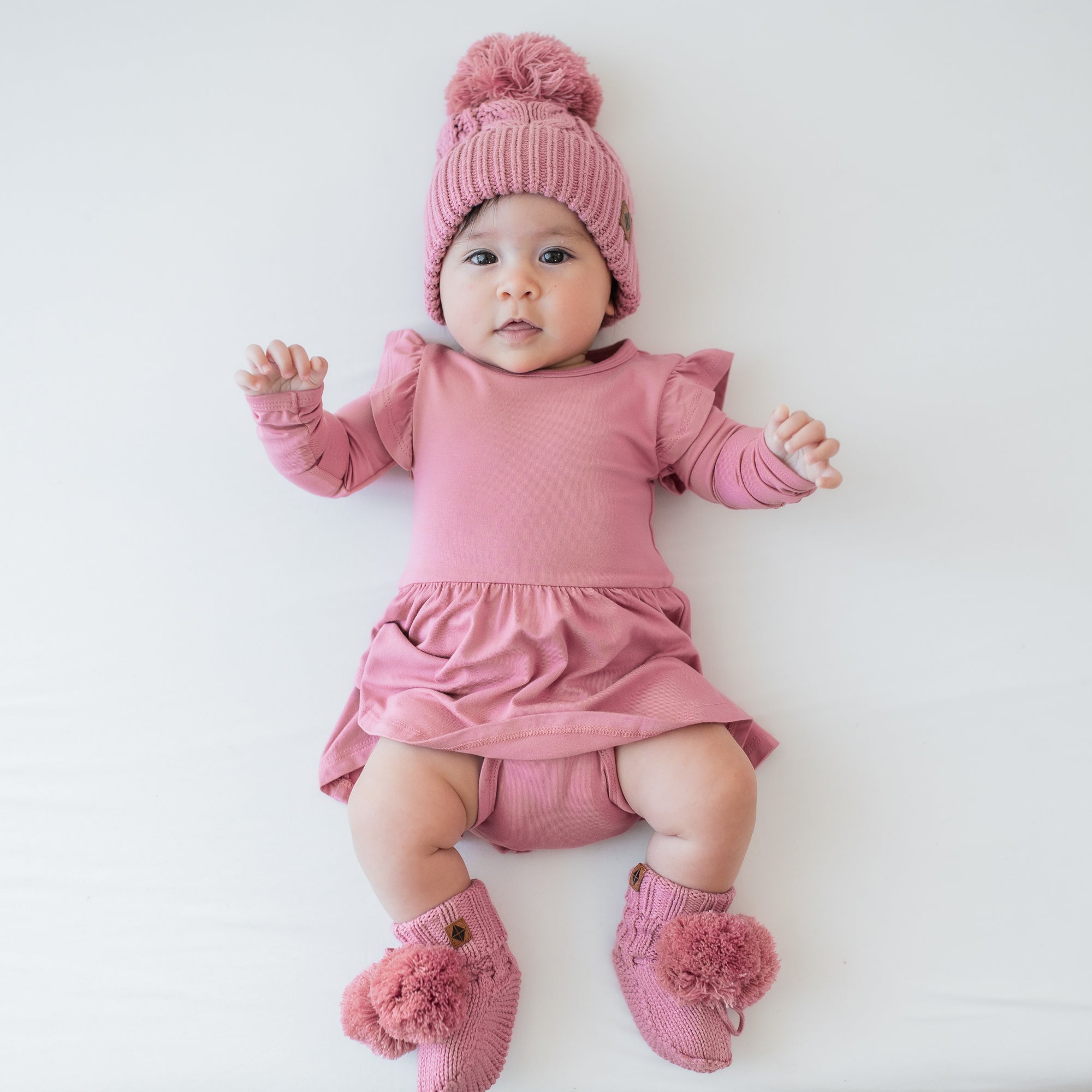 Infant model shown wearing the Chunky Cable Knit Beanie in Apple Blossom paired with a matching long sleeve twirl bodysuit dress and knit booties
