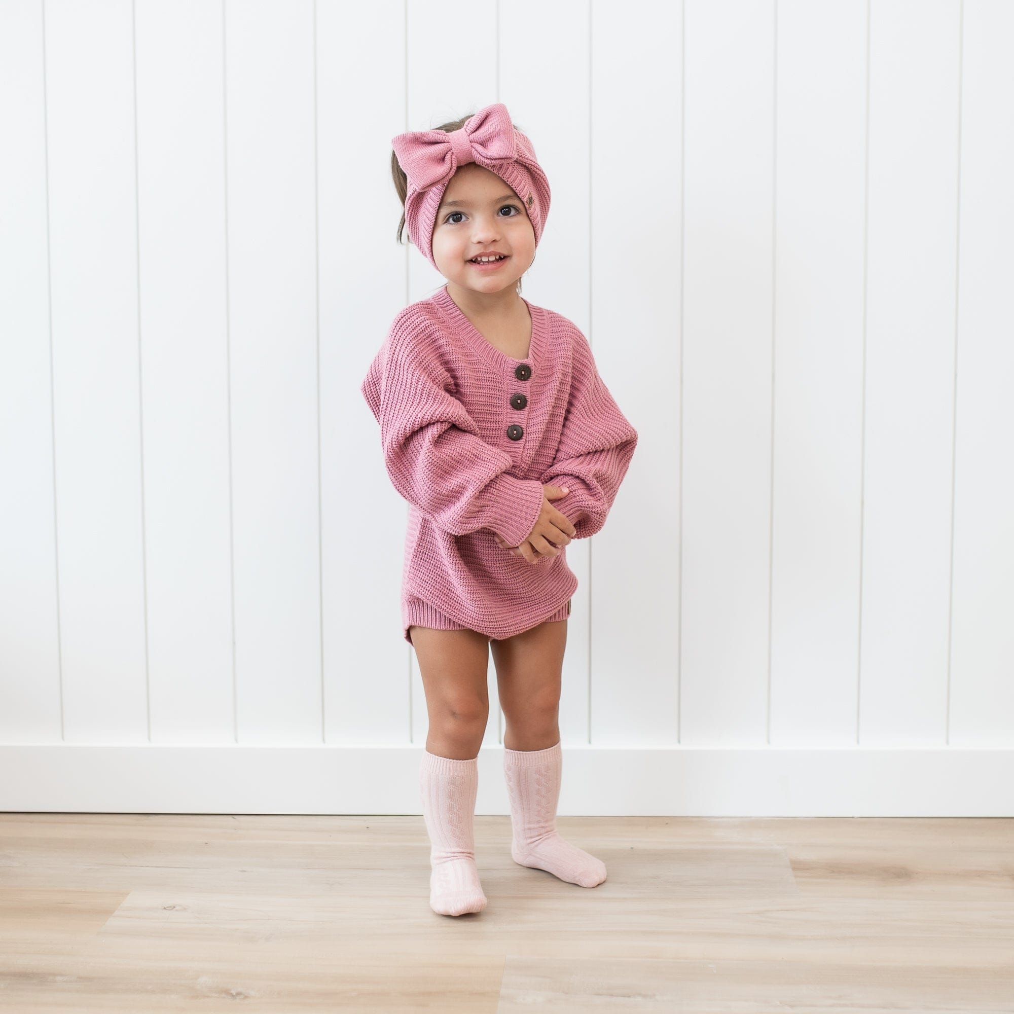 Toddler standing in front of a white paneled wall wearing the Chunky Knit Bow Headband in Apple Blossom and matching chunky knit romper