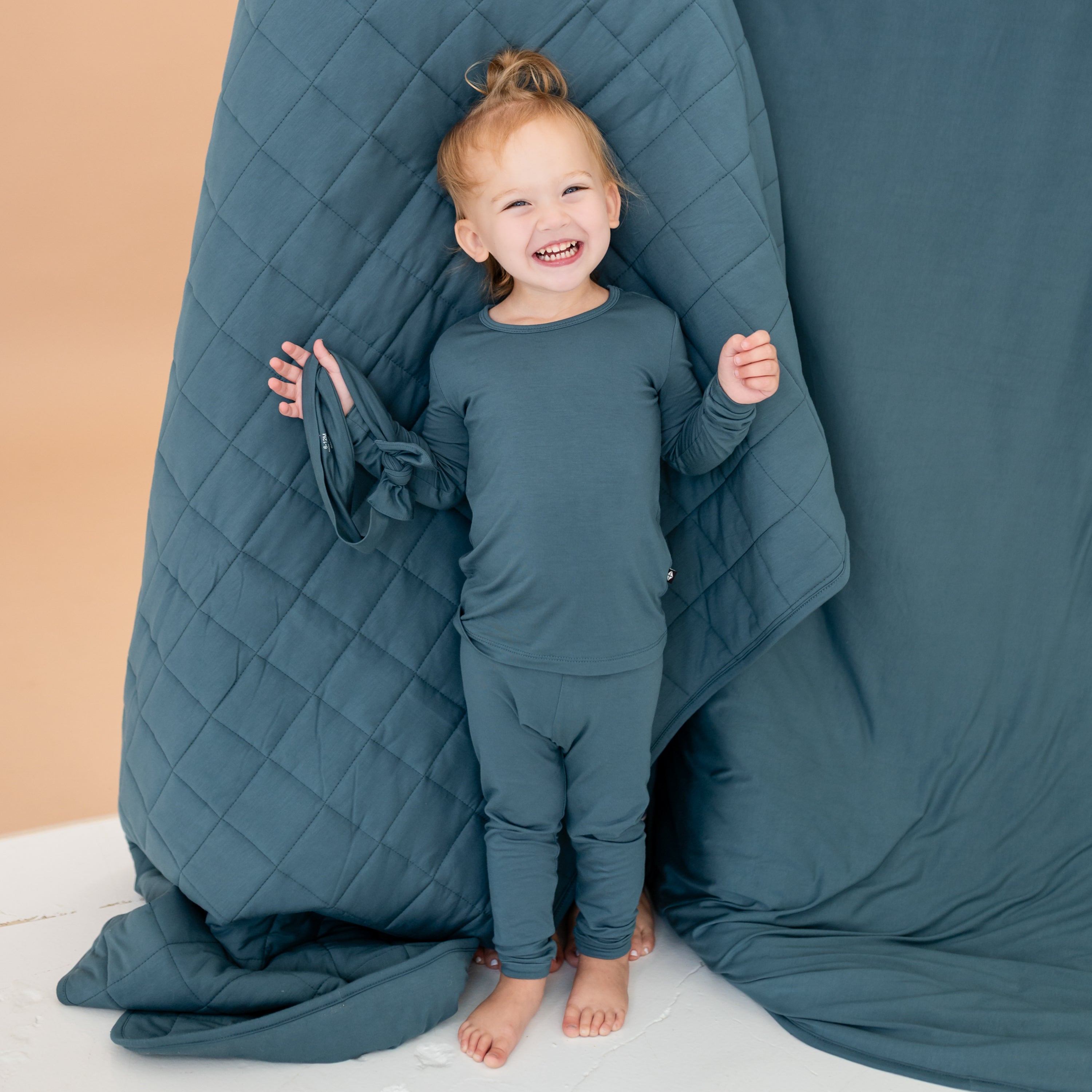 Smiling toddler standing in front of an Atlantic blanket holding a knotted bow headband wearing the Long Sleeve Pajamas in Atlantic