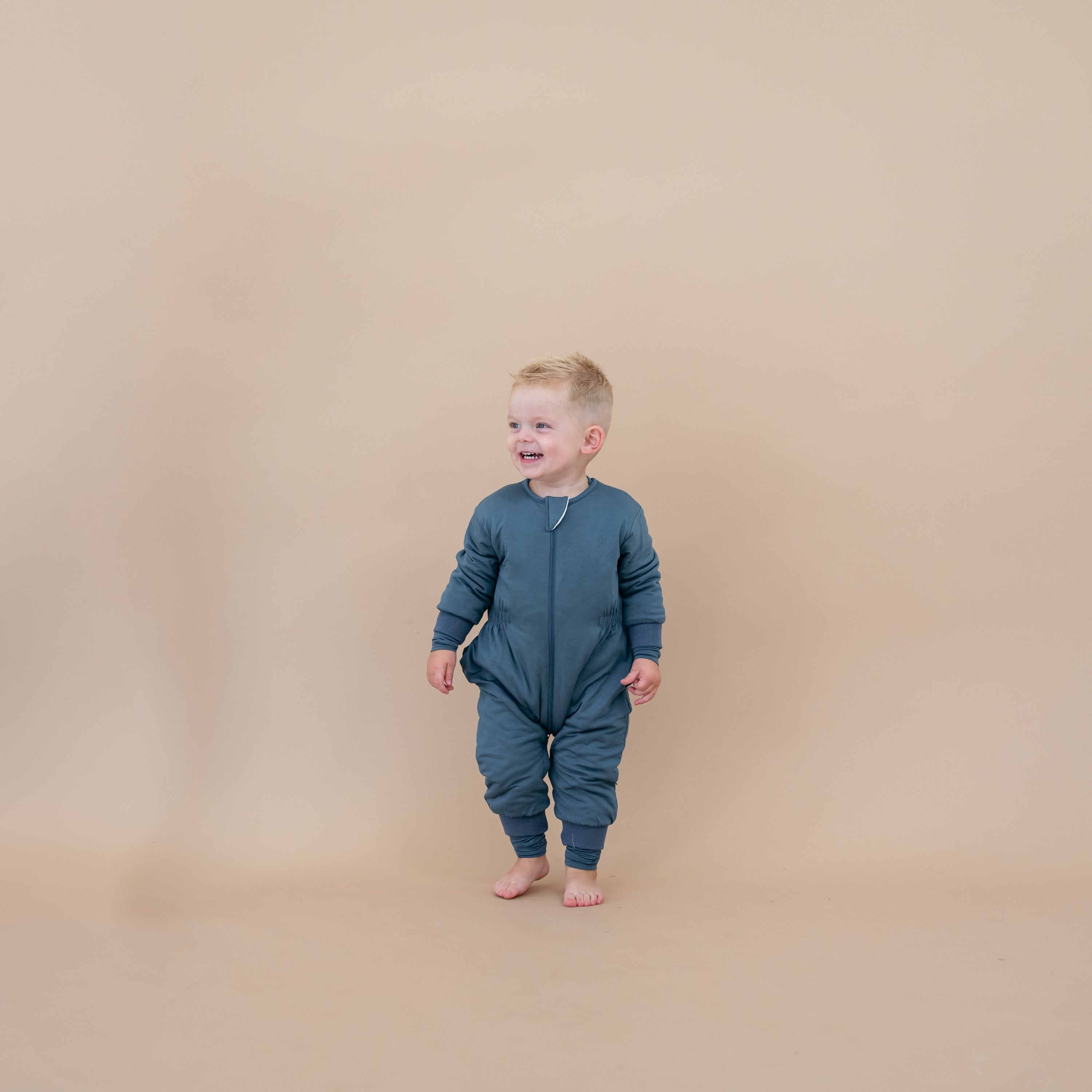 Toddler walking wearing the Cozy Playsuit in Atlantic in front of a light taupe backdrop