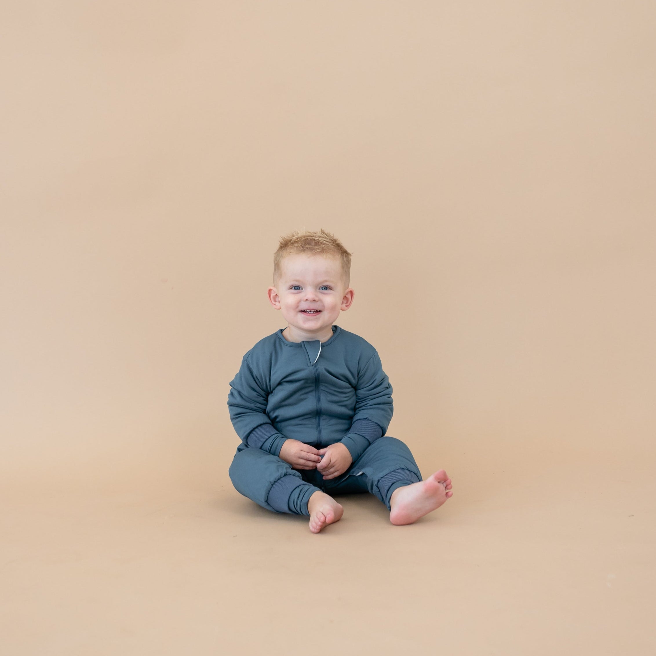 Toddler smiling sitting wearing the Cozy Playsuit in Atlantic in front of a light taupe backdrop