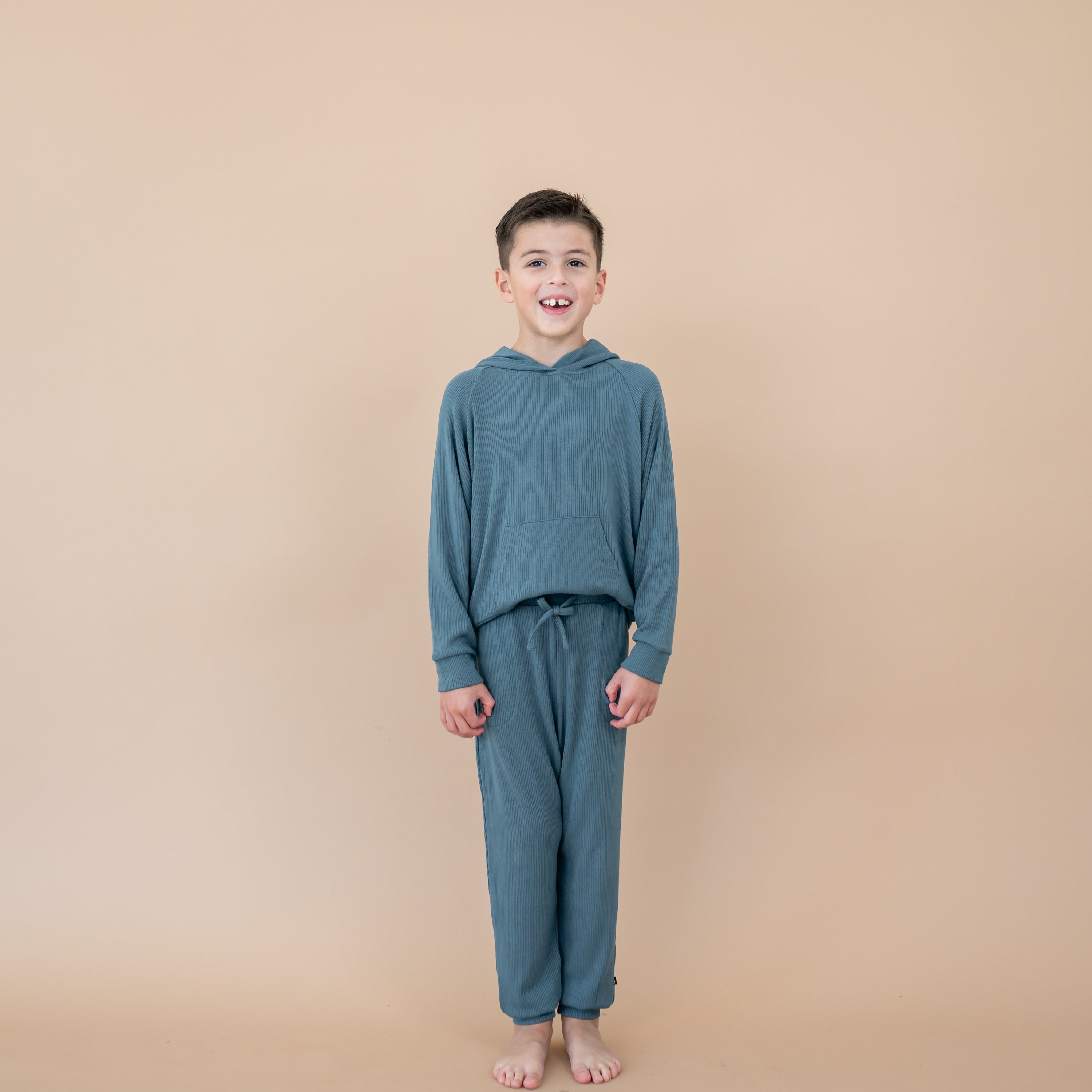 Smiling boy model wearing the Ribbed Jogger Pant in Atlantic paired with the matching ribbed hoodie in front of a taupe background