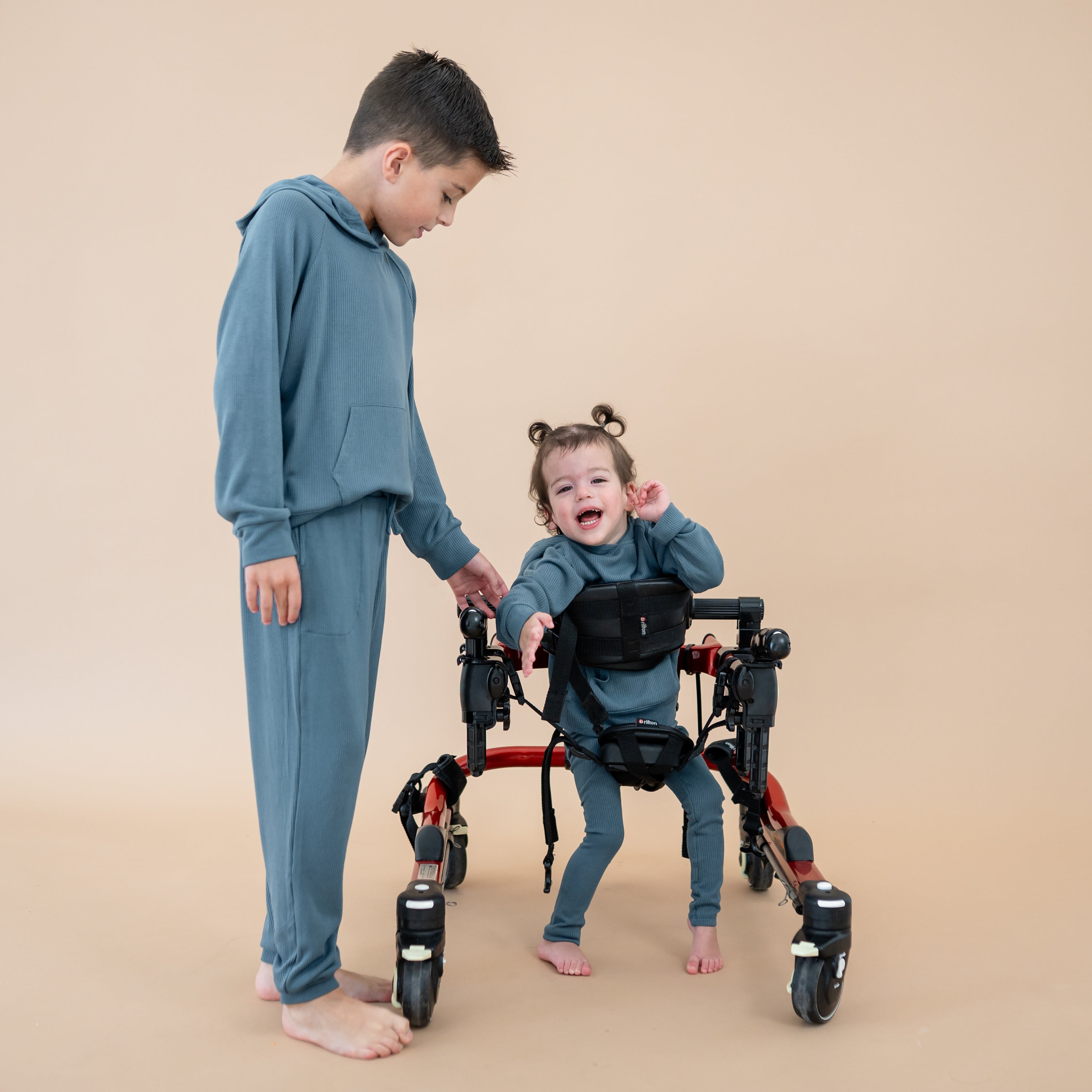 Brother wearing the Ribbed Jogger Pant in Atlantic and matching Ribbed hoodie standing beside his sister in her walker who is wearing the Ribbed Hoodie in Atlantic and matching ribbed leggings in front of a taupe background