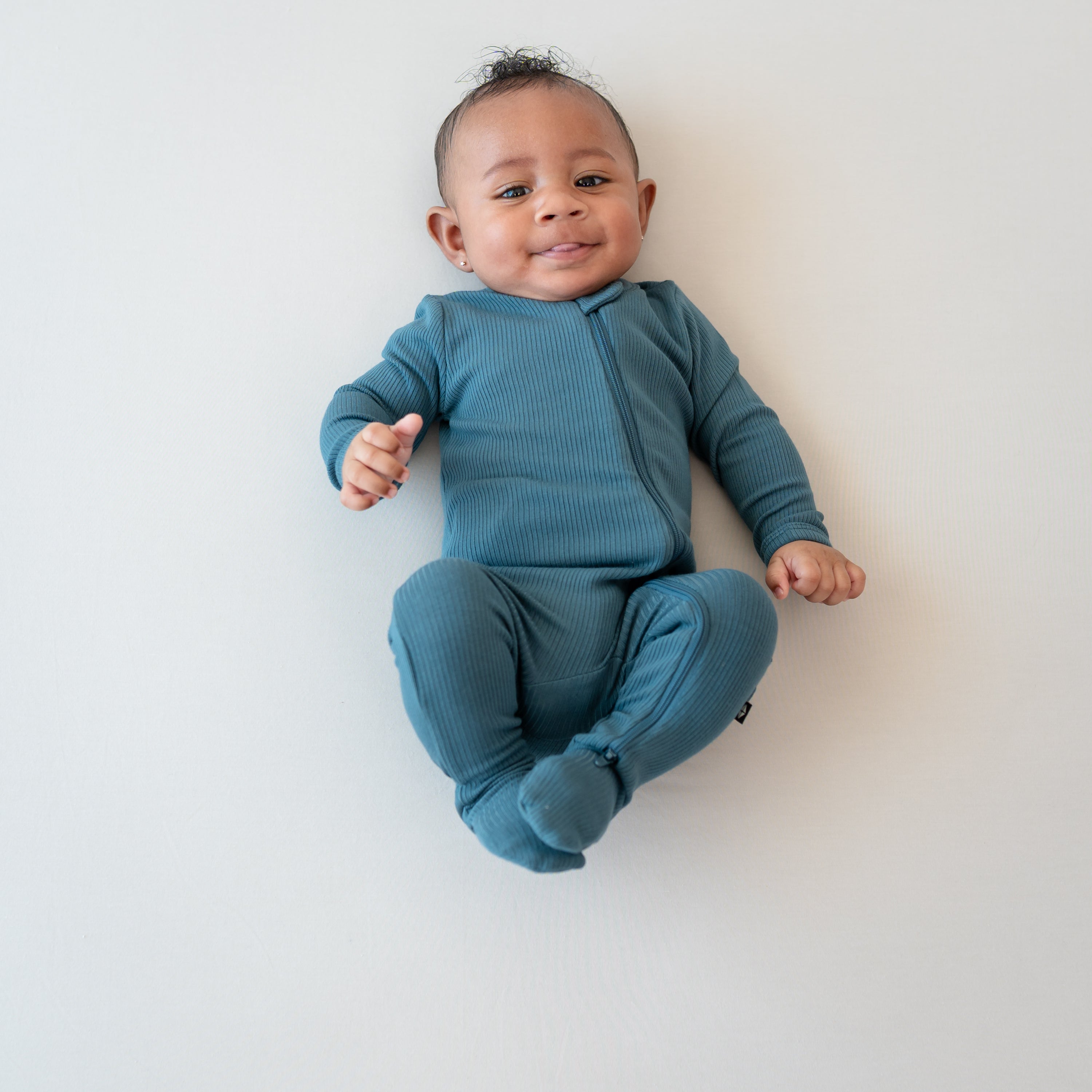 Smiling infant sticking their tongue out wearing the Ribbed Zipper Footie in Atlantic on a bisque blanket