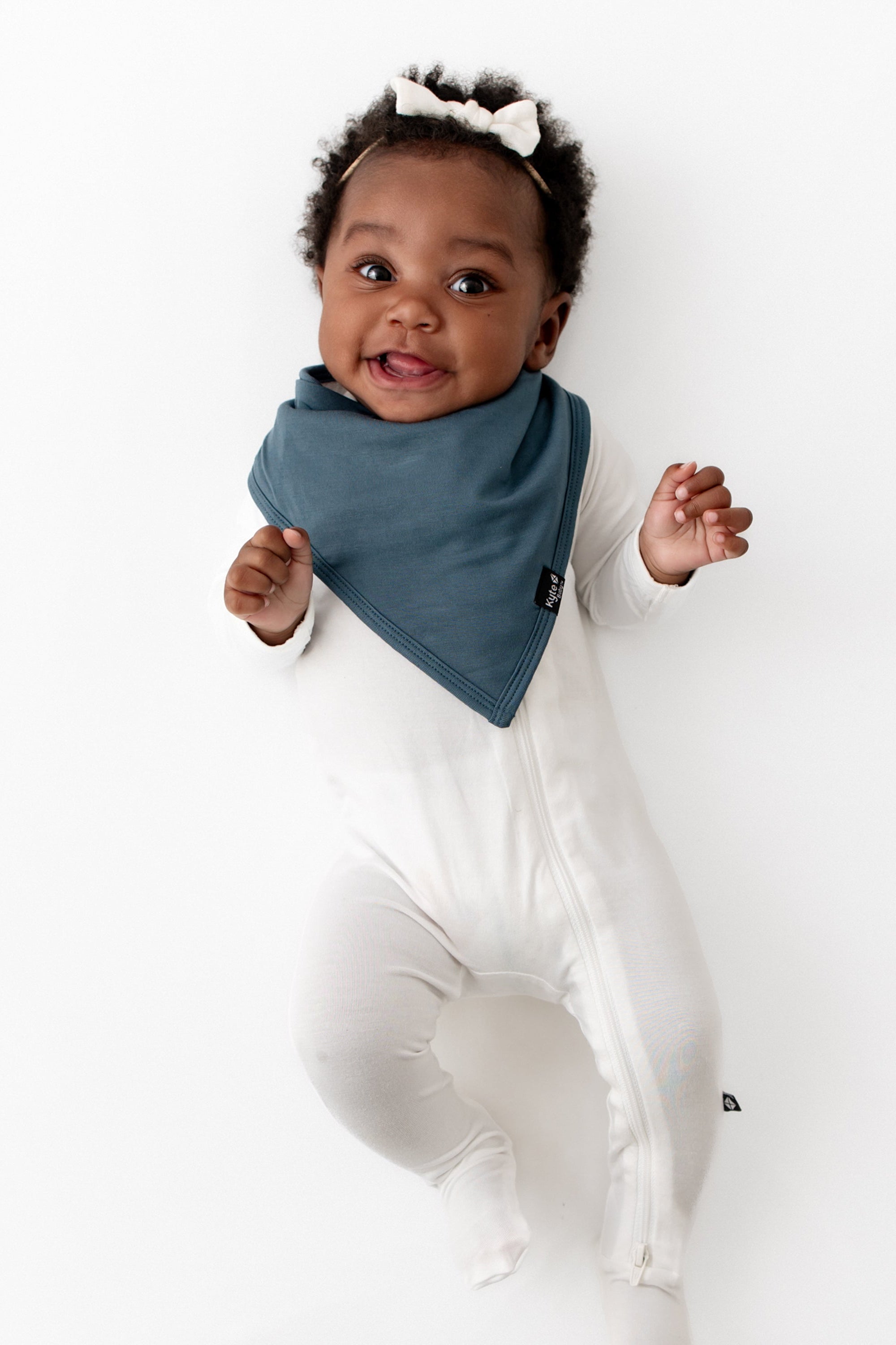 Infant girl laying down wearing the Bib in Atlantic paired with a Cloud Zippered Footie and Cloud Tiny Nylon Bow