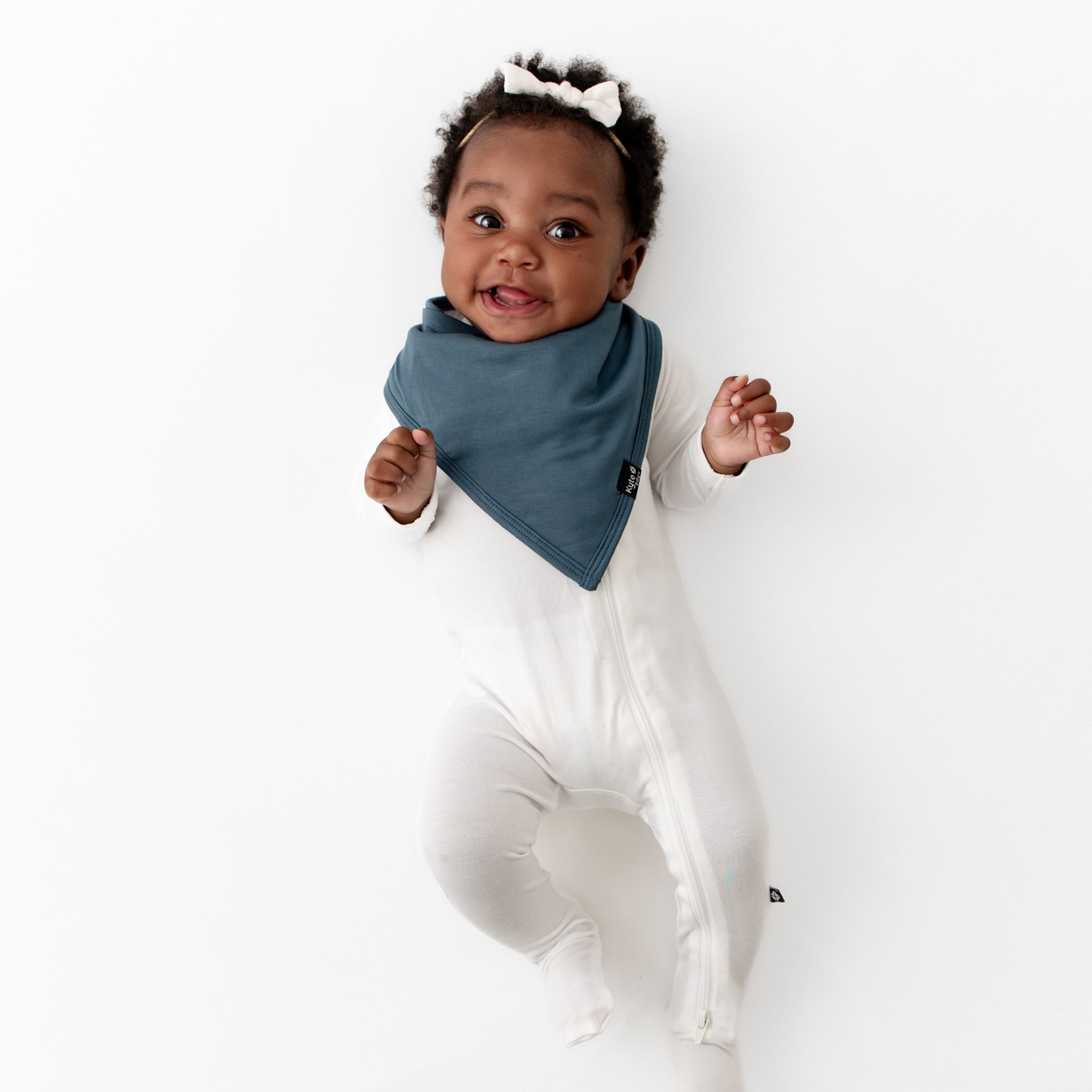 Infant girl laying down wearing the Bib in Atlantic paired with a Cloud Zippered Footie and Cloud Tiny Nylon Bow