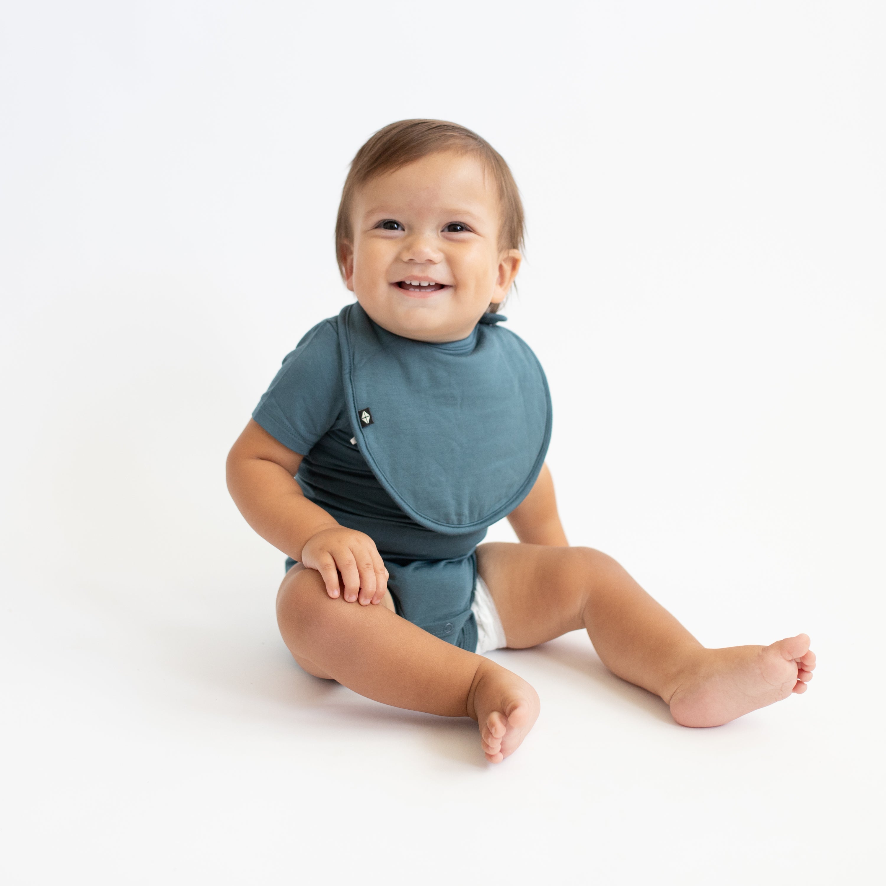Smiling infant sitting down wearing the Cushy Bib in Atlantic with matching short sleeve bodysuit