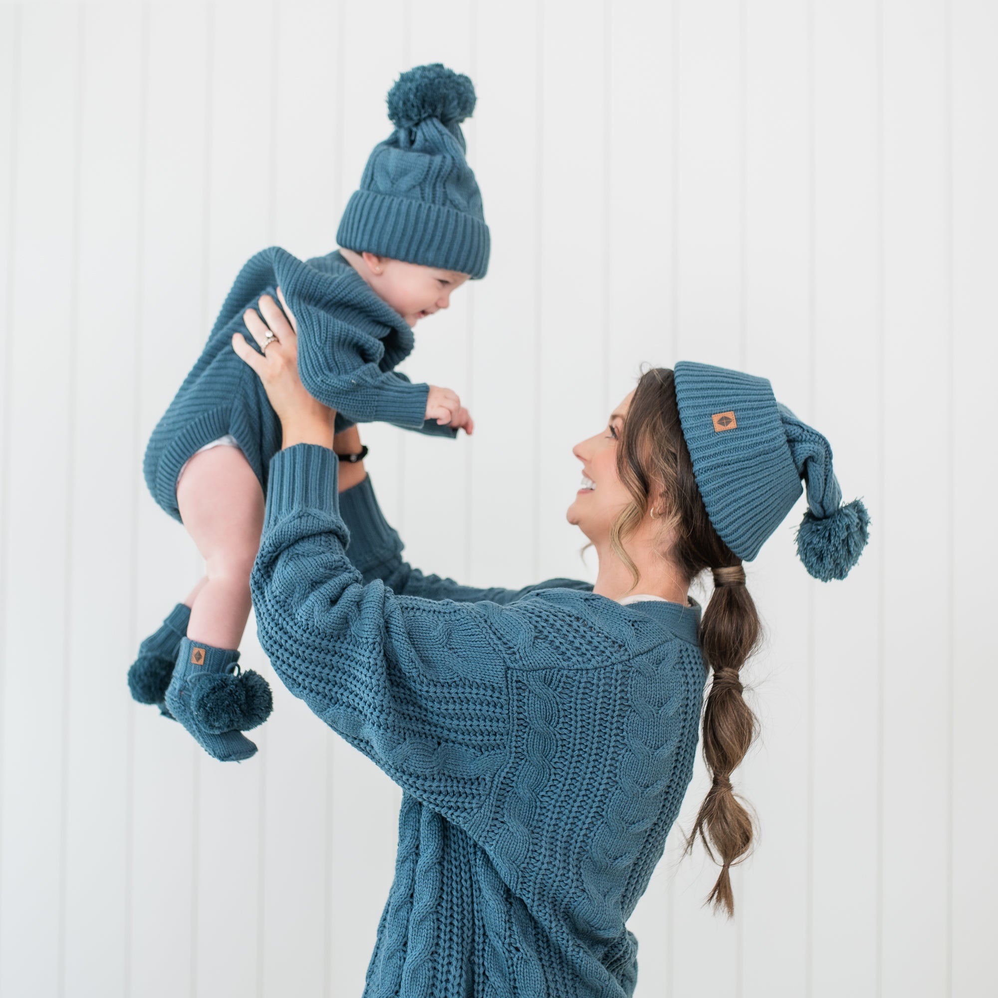 Mother holding toddler in the air both wearing chunky knit items in the color Atlantic