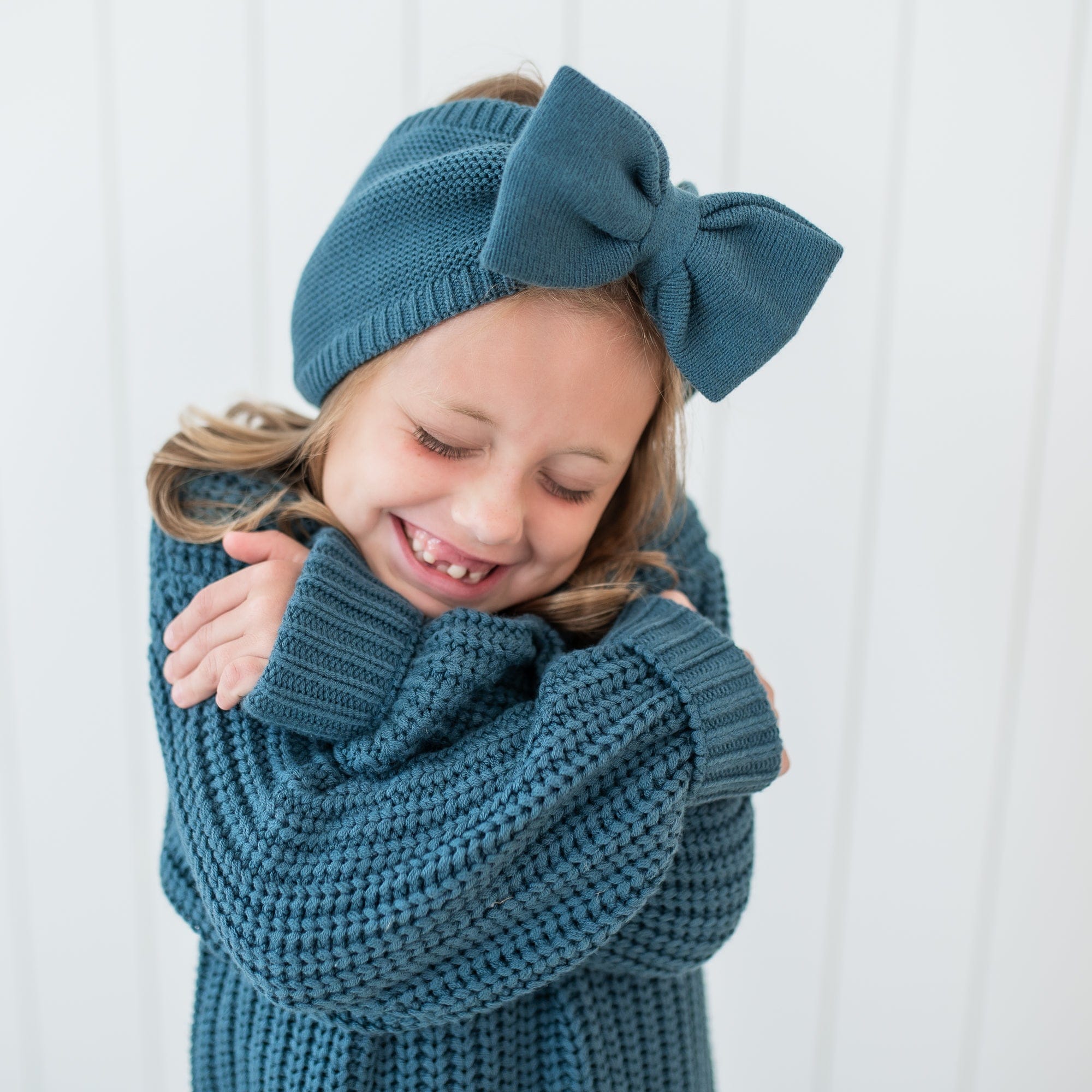 Young girl hugging herself wearing the Chunky Knit Bow Headband in Atlantic and matching Chunky Knit Sweater