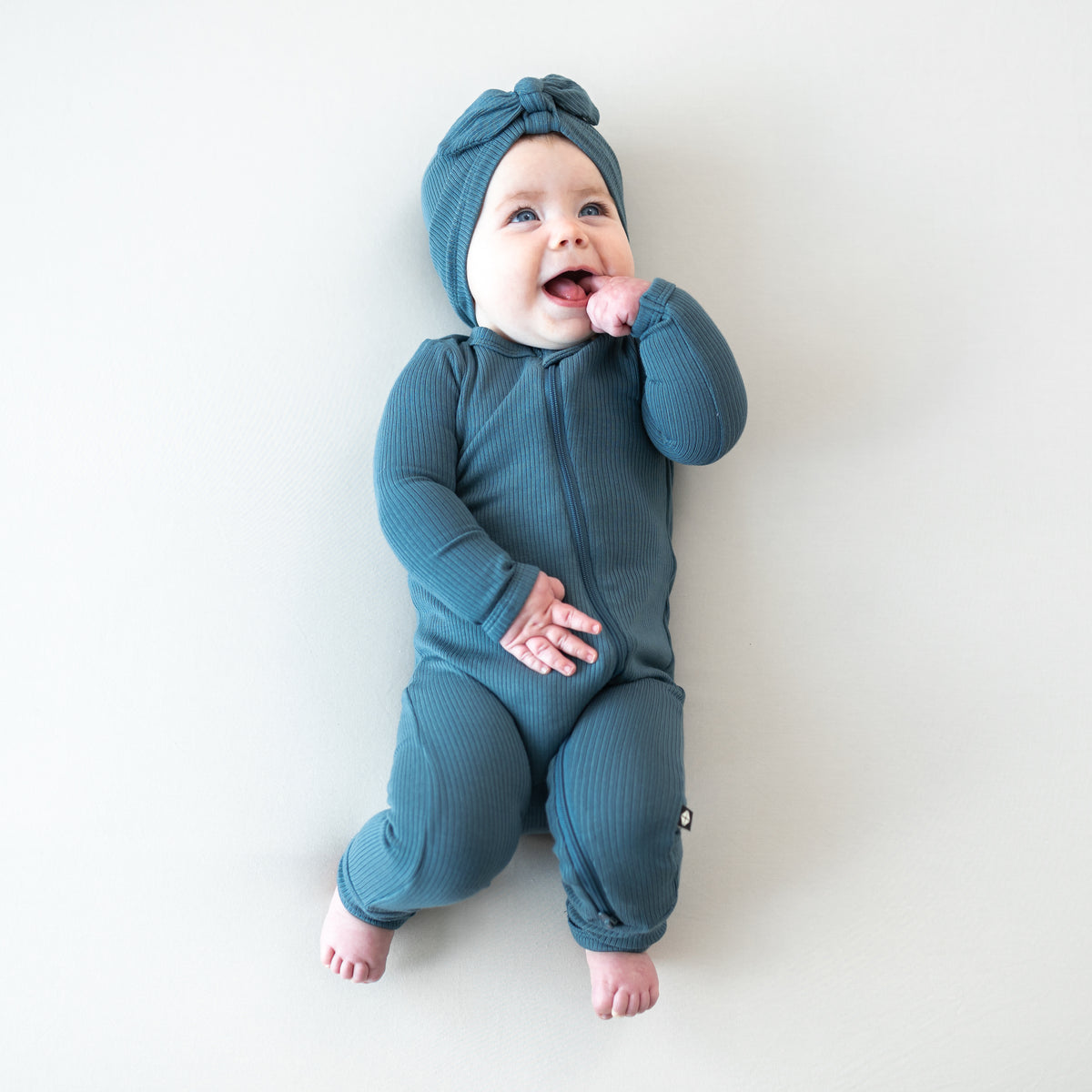 Smiling infant laying down on a light background wearing the Ribbed Headwrap in Atlantic paired with a matching ribbed zipper romper
