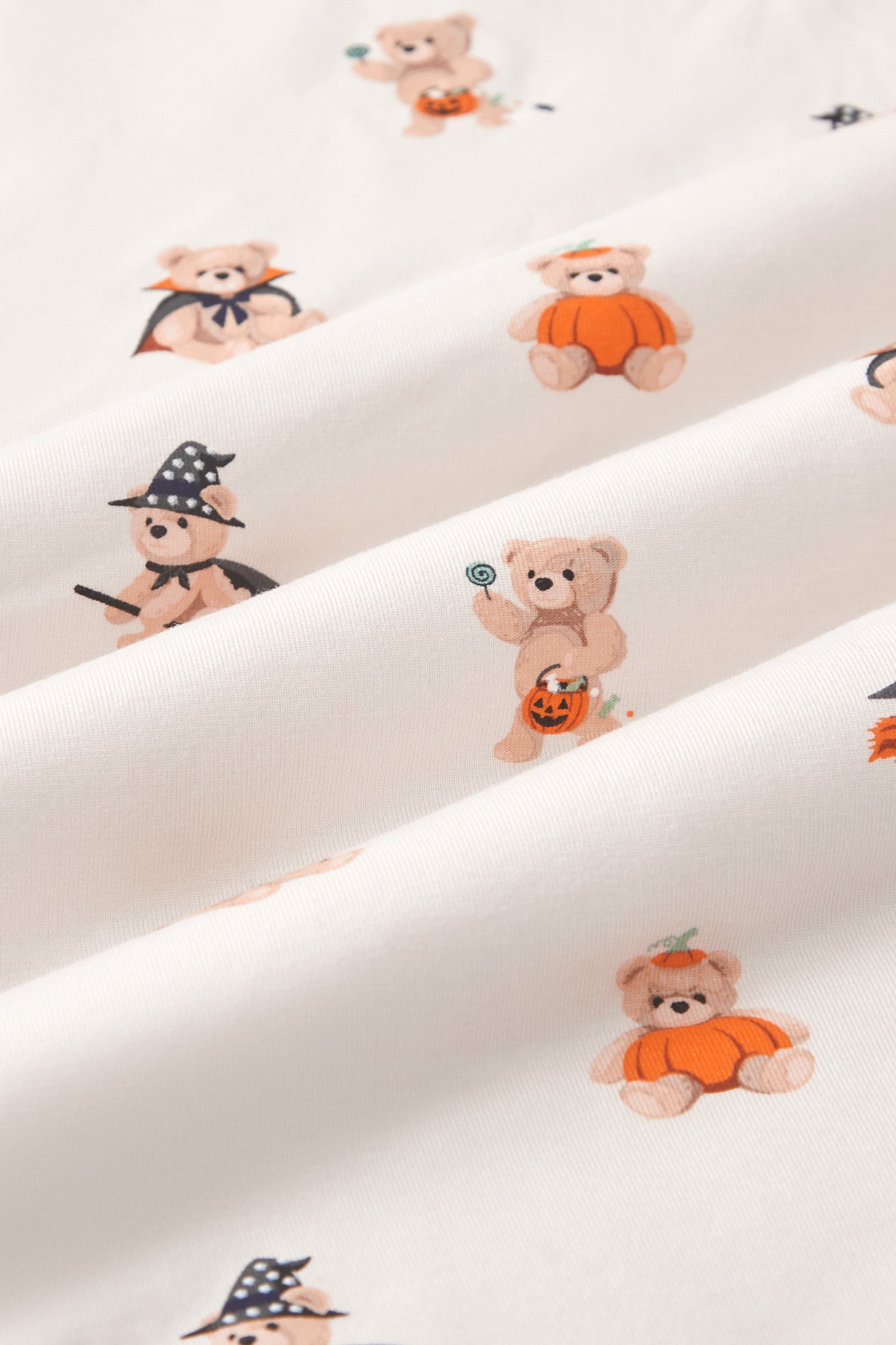 A repeating, all-over print featuring: a light brown teddy bear with white fangs, wearing a black and orange vampire cape and a purple bow around its neck;a teddy bear holding a lollipop in one hand and a jack o’ lantern basket with candy falling out in the other;a teddy bear sitting inside an orange pumpkin and wearing the pumpkin top on its head; a teddy bear riding a black and orange broom,wearing a starry black witch’s hat and black cape around its neck, over an off-white background color called Vanilla