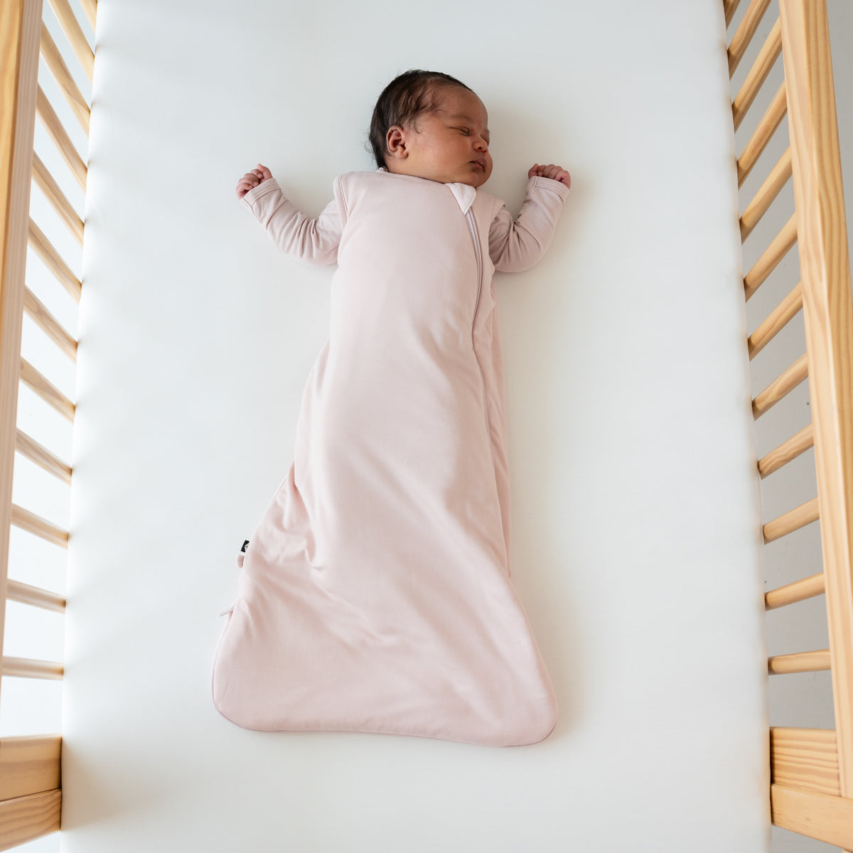 Baby sleeping in a wooden crib while wearing a Kyte Baby Sleep Bag in Blush 1.0 TOG.