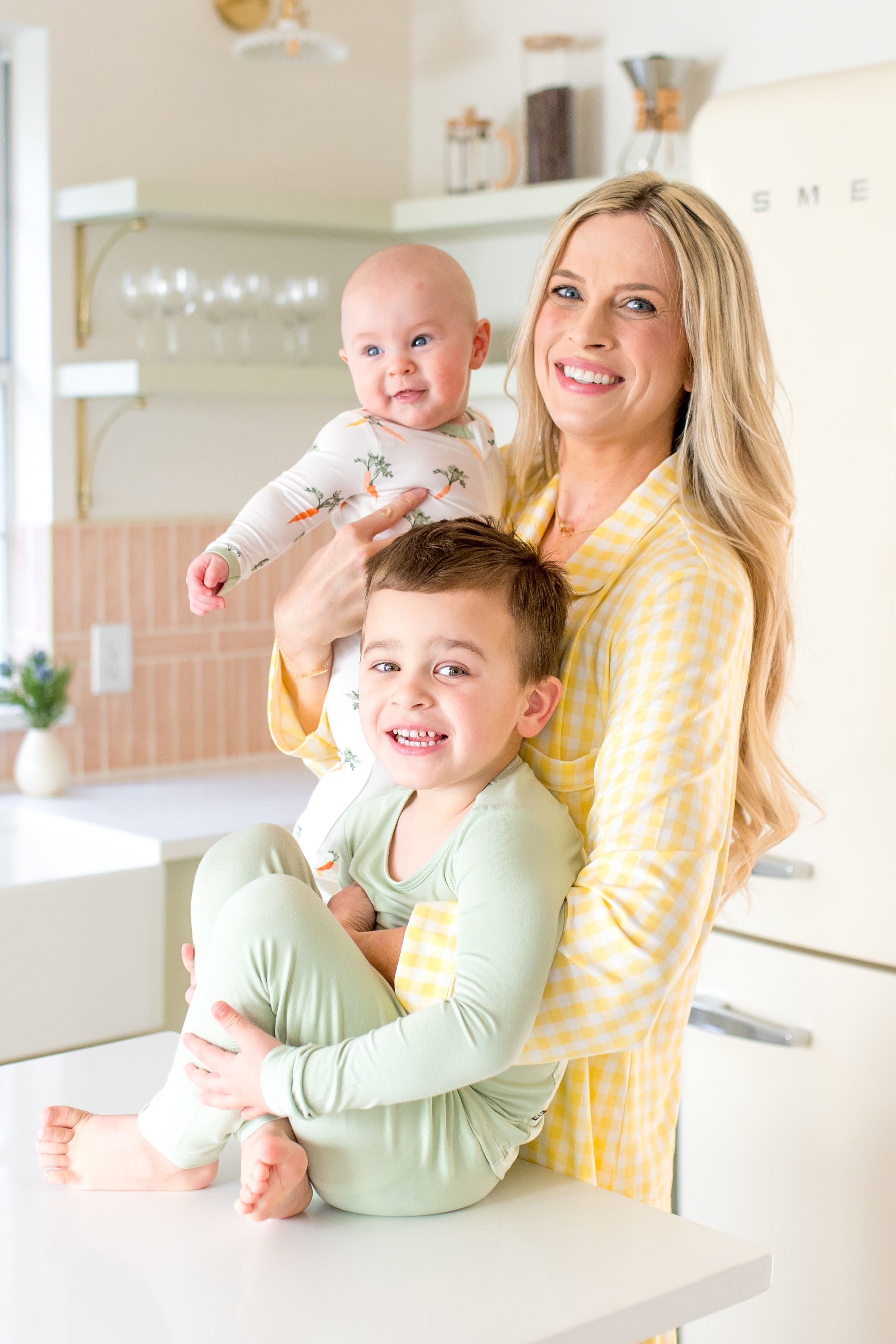Woman holding a baby and a toddler in a kitchen with a Smeg refrigerator.
