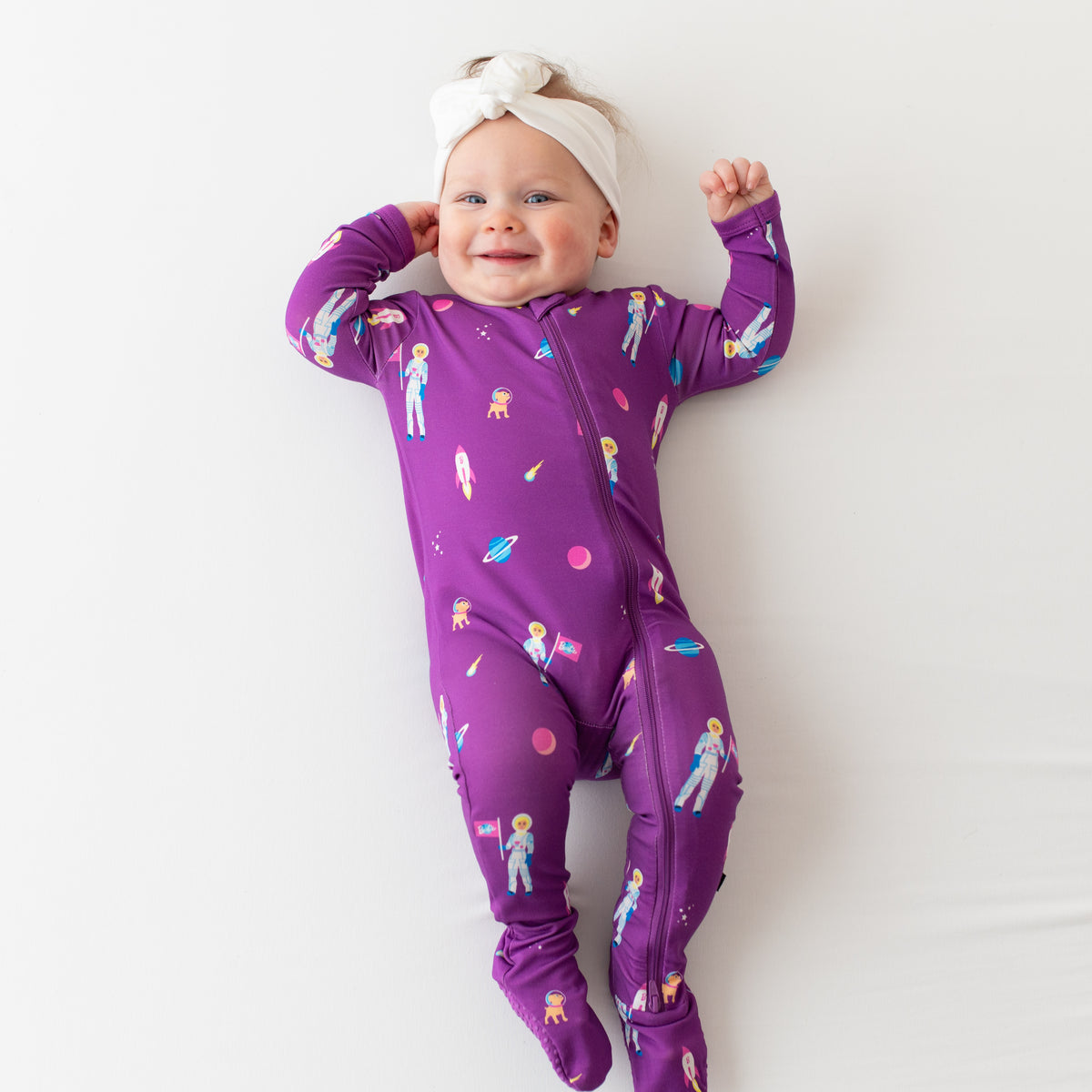 Infant wearing Kyte Baby Zippered Footie in Barbie™ Astronaut and Cloud Knotted Bow