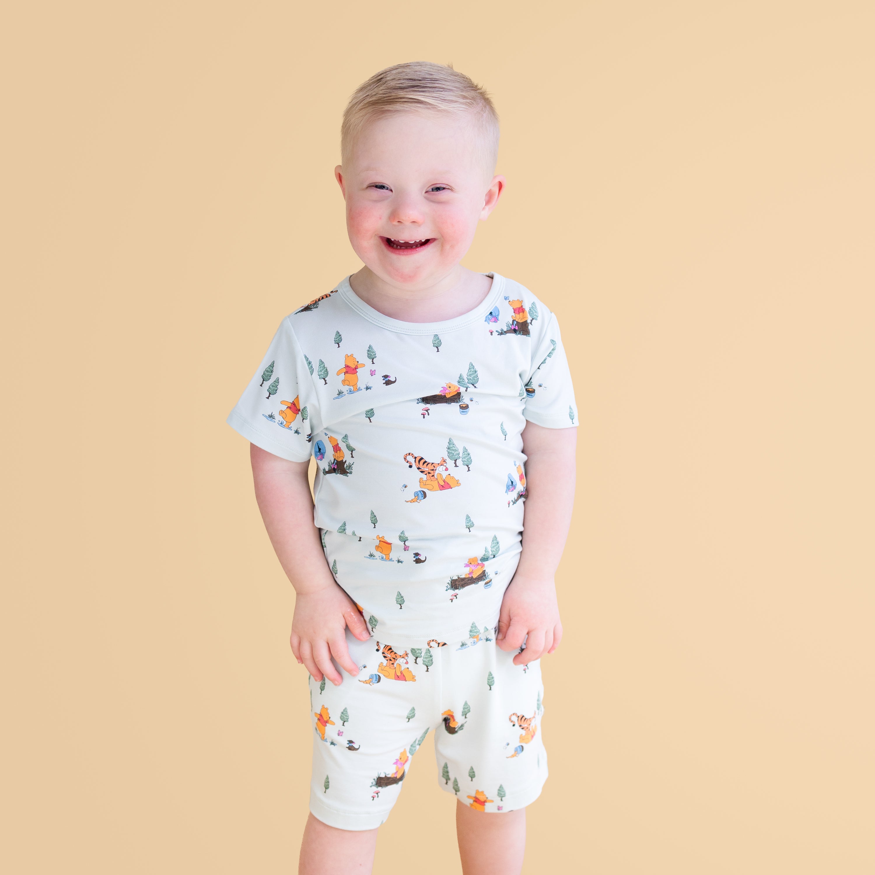 Smiling boy modeling the Toddler Basic Tee in Winnie the Pooh Hundred Acre Wood paired with matching shorts standing in front of a mustard yellow background