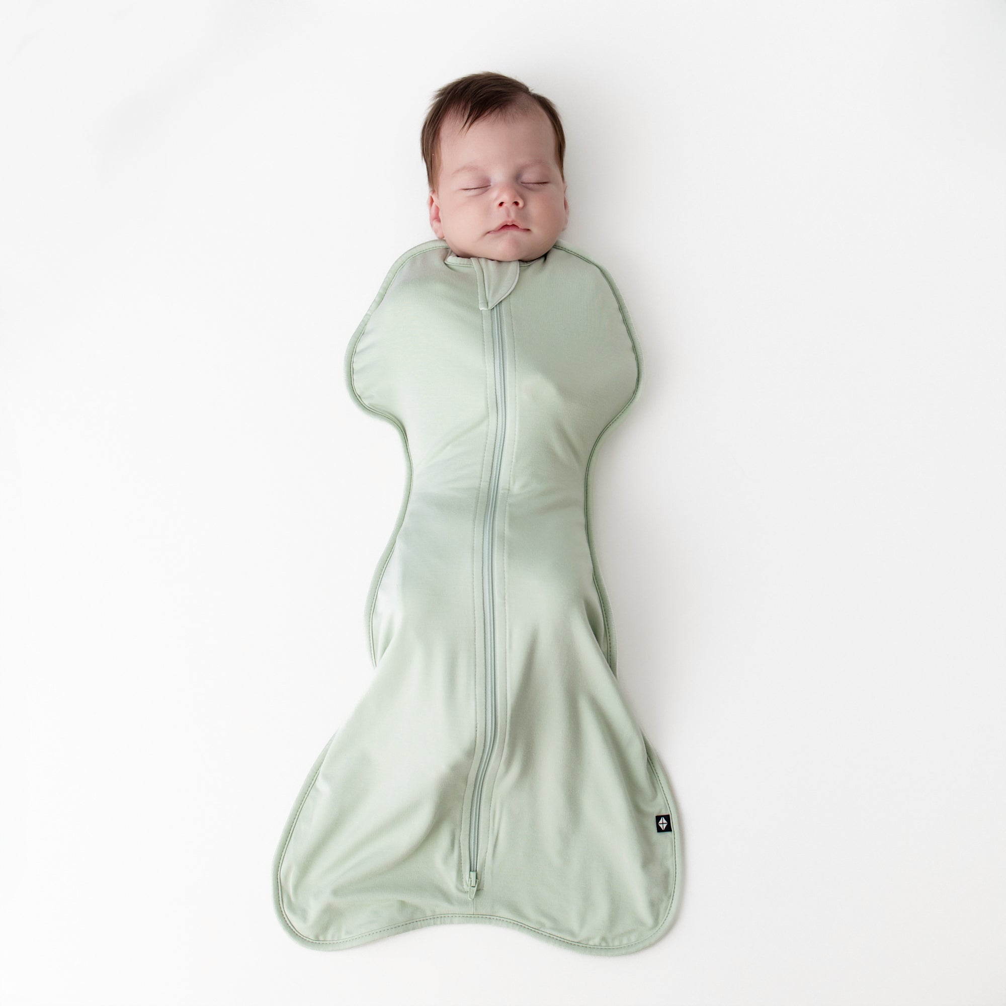 Sleeping newborn wearing the Swaddle Bag in Basil 0.5 laying on a white surface