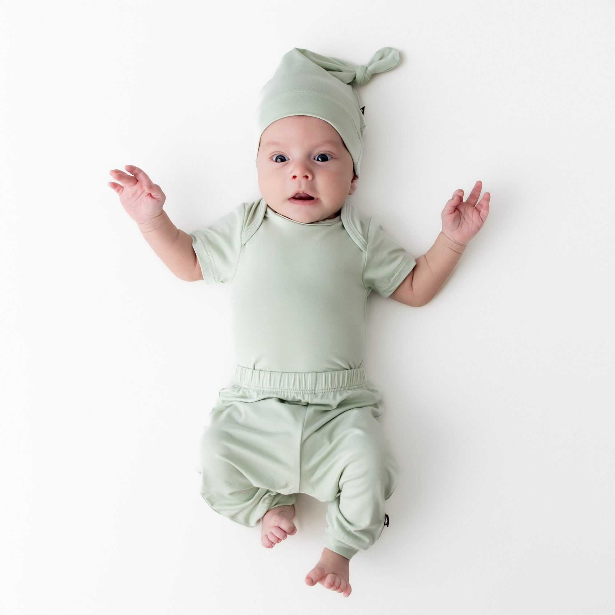 Newborn laying on a cream surface wearing the Knotted Cap in Basil with matching short sleeve bodysuit and pants