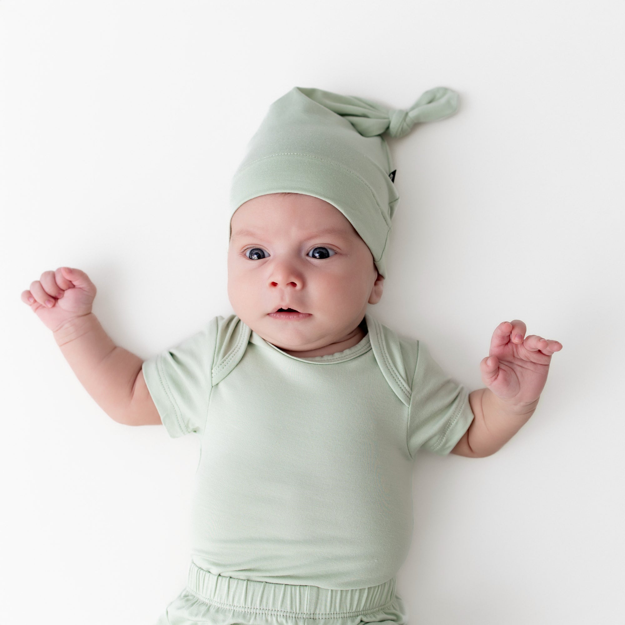 Newborn wearing the Knotted Cap in Basil with matching short sleeve bodysuit laying on a cream surface