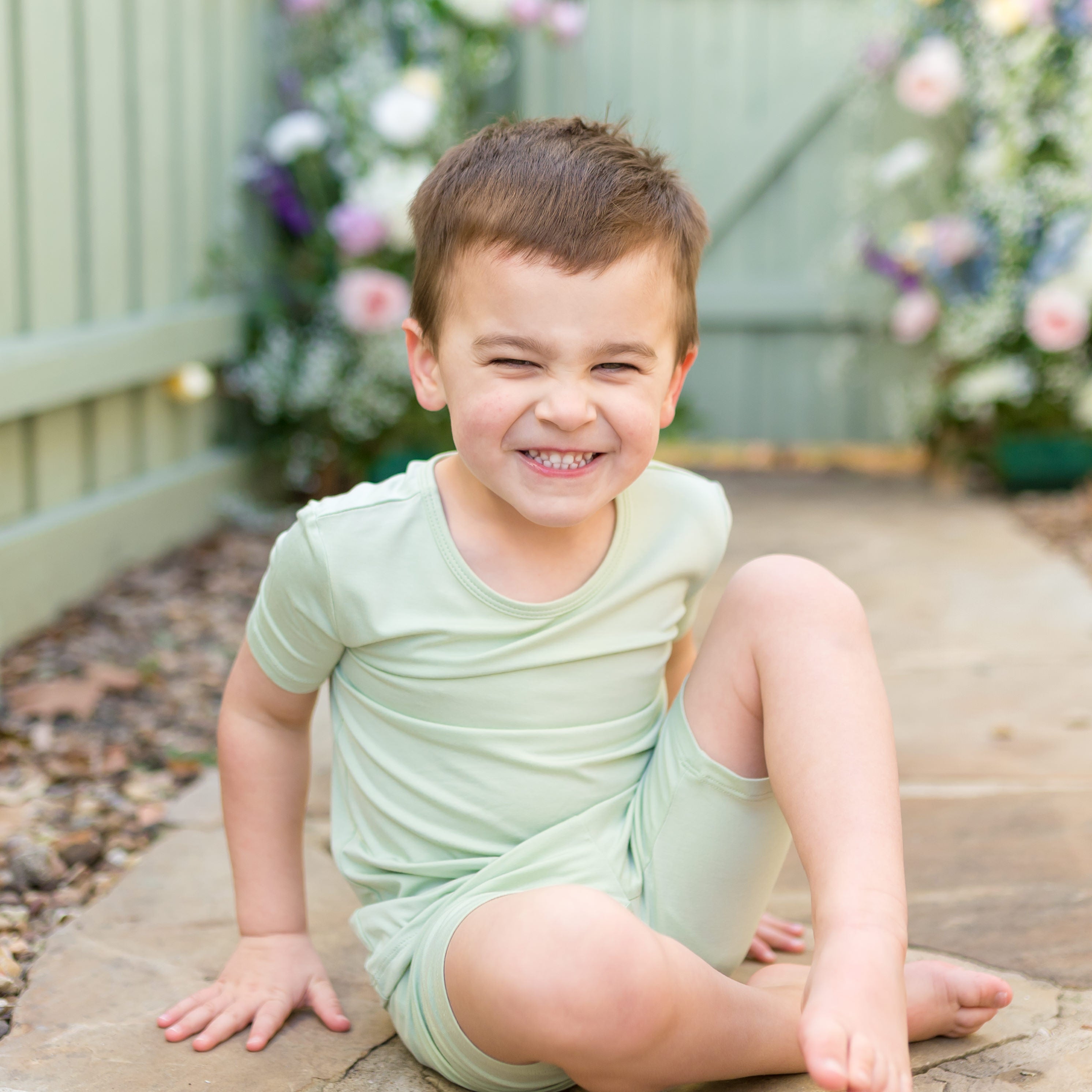 Young smiling boy sitting on a stone path wearing the Short Sleeve Pajamas in Basil