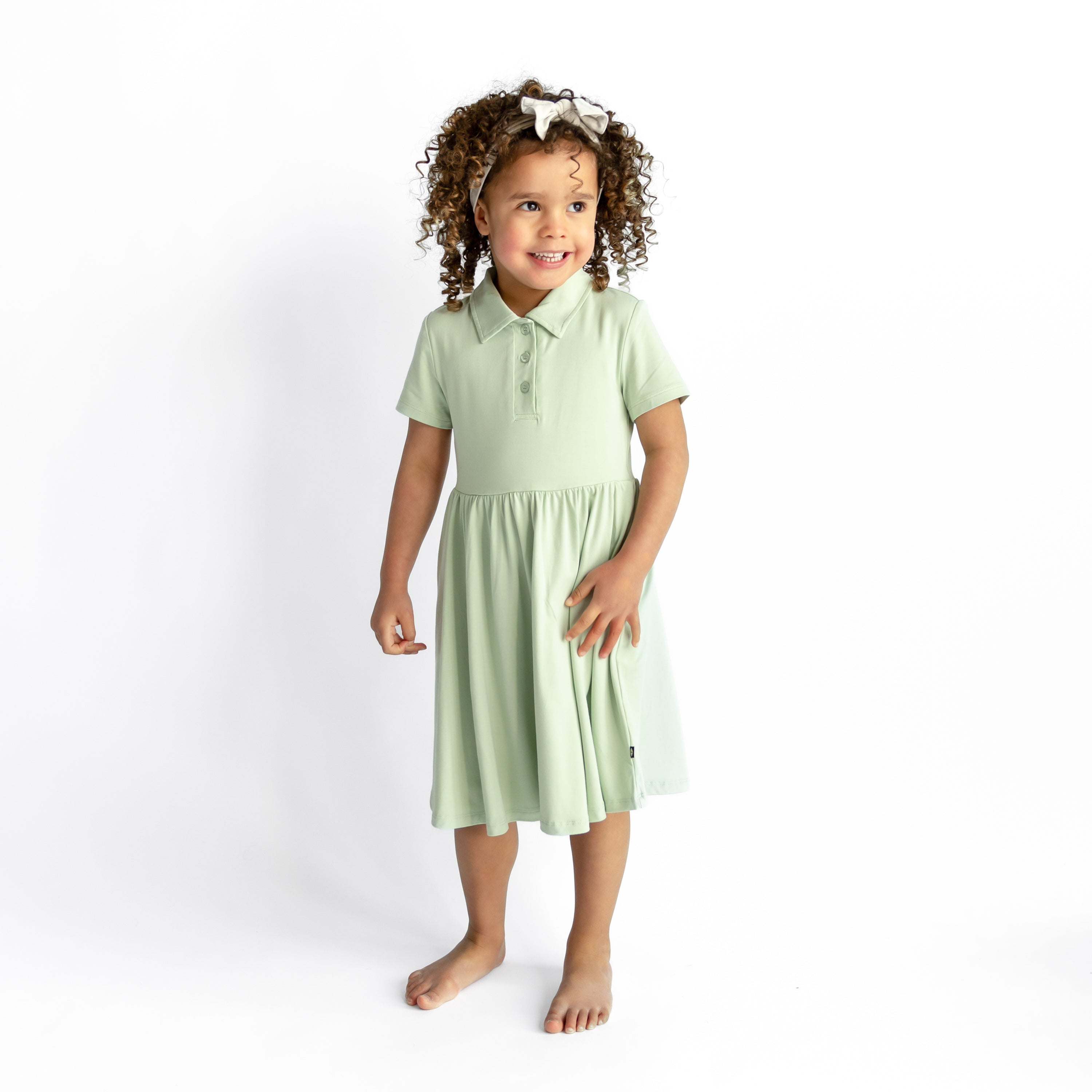 Young girl smiling wearing the Polo Dress in Basil standing in front of a white background