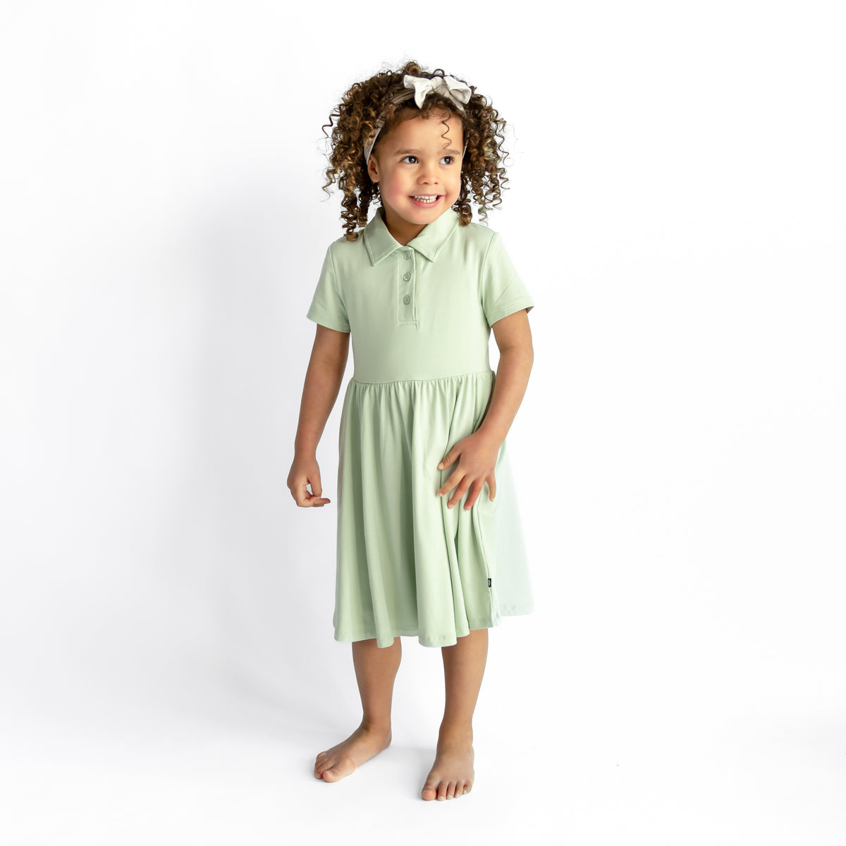 Young girl smiling wearing the Polo Dress in Basil standing in front of a white background