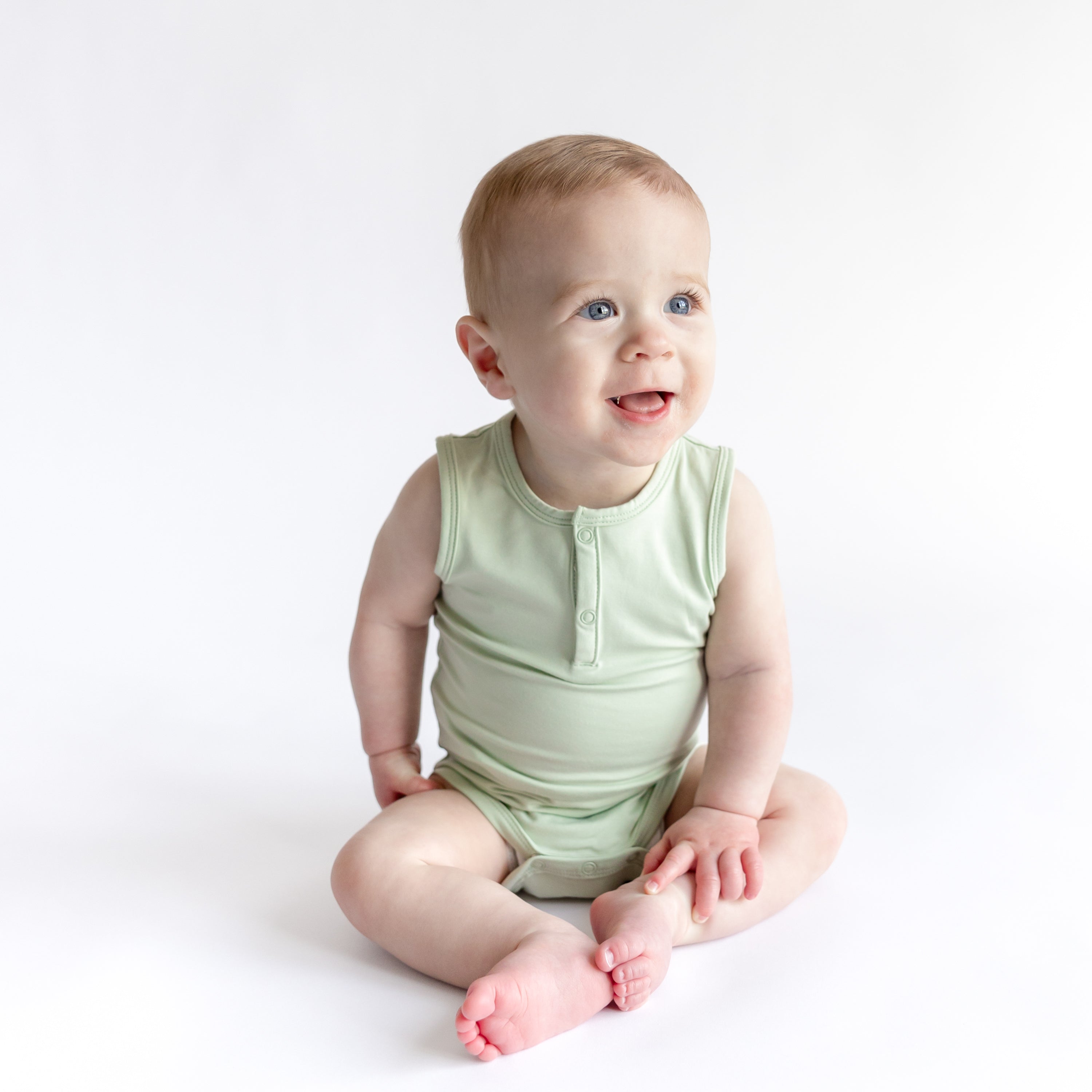 Young toddler sitting on the floor wearing the Sleeveless Bodysuit in Basil in front of a white background