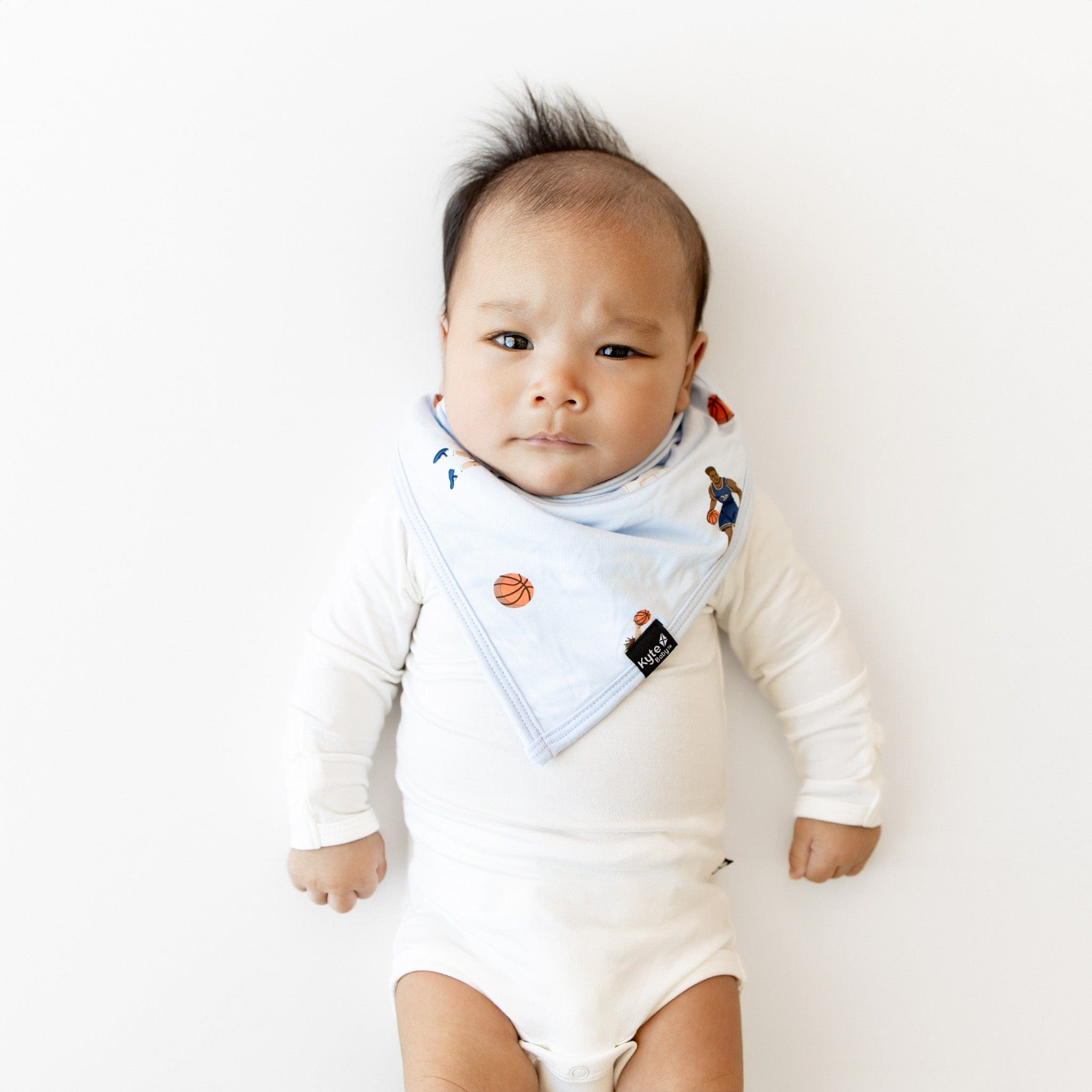 Infant laying on a white surface wearing the Bib in Basketball overtop a white long sleeve bodysuit
