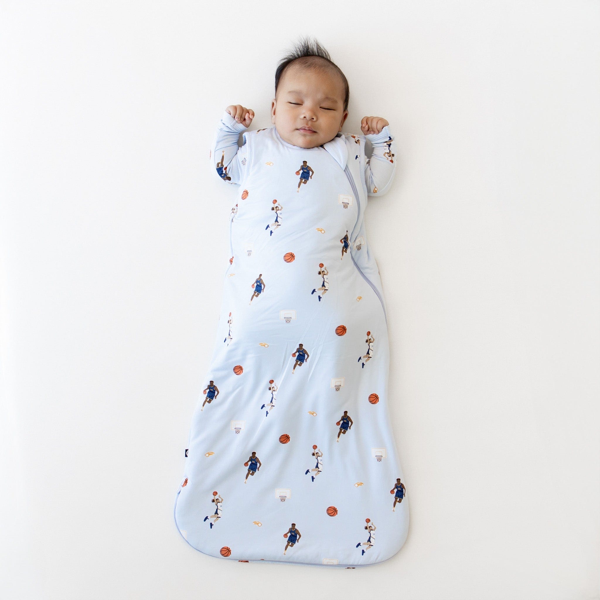 Sleeping infant laying on a light neutral surface wearing the Sleep Bag in Basketball 1.0 and matching romper